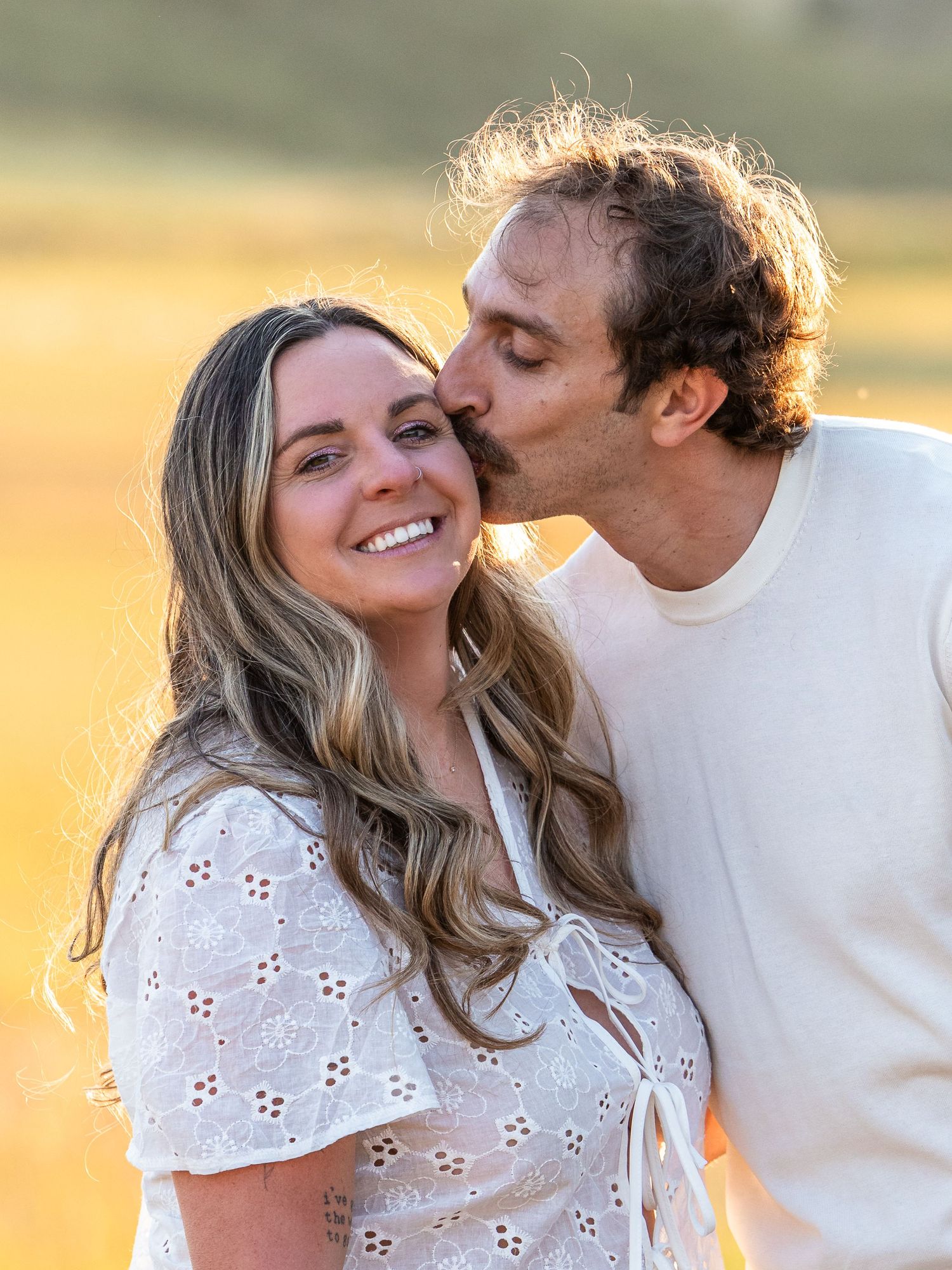 A couple shares a tender moment outdoors at golden hour, man kissing woman's cheek as she smiles warmly.