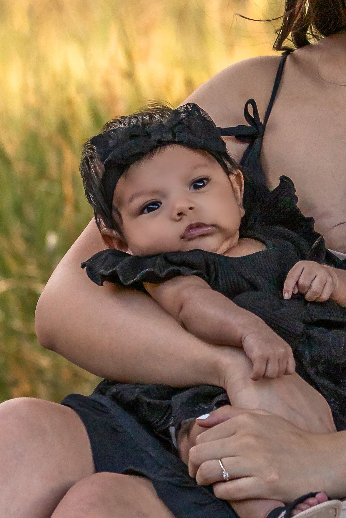 A mother in black dress holds her newborn baby girl wearing a black outfit and headband outdoors in a golden field.