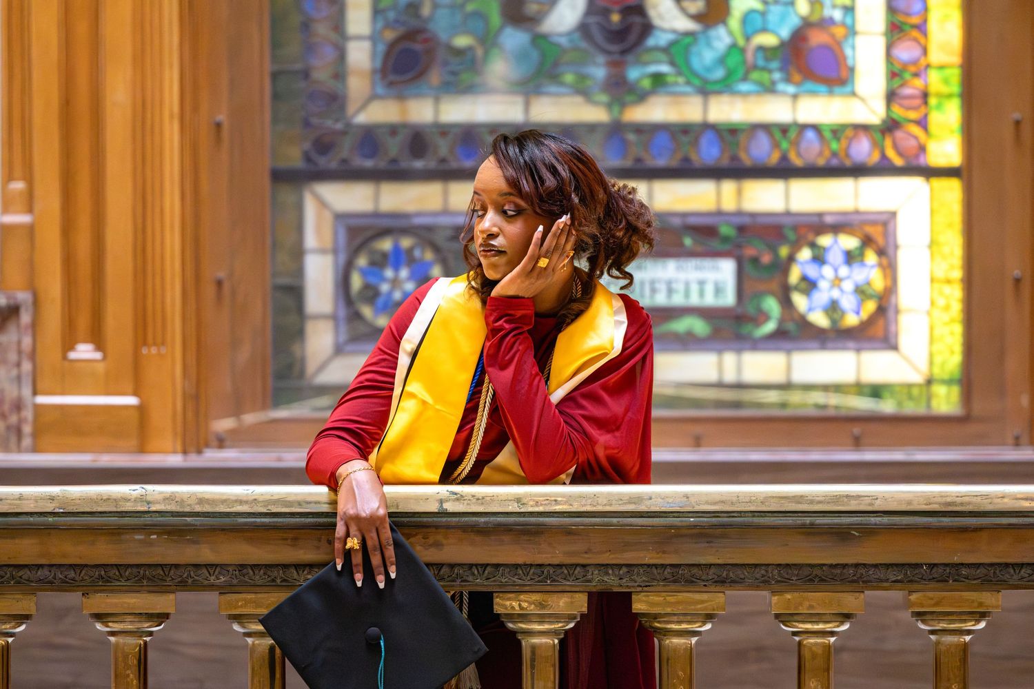 Graduate in red and yellow regalia holds cap, posing by ornate railing with colorful stained glass window behind.