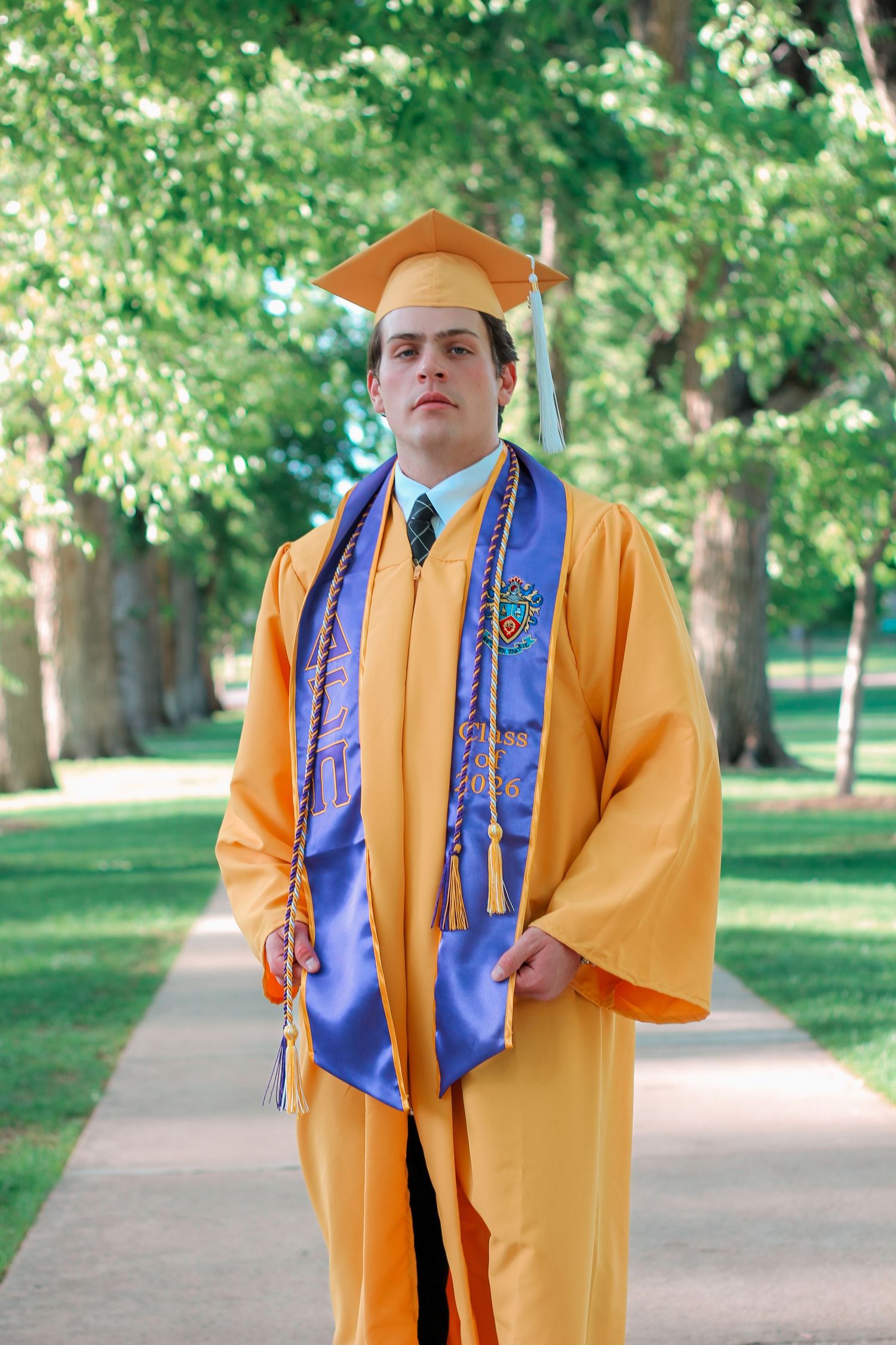 Young man in orange graduation cap and gown with blue honor stole poses on tree-lined campus walkway.