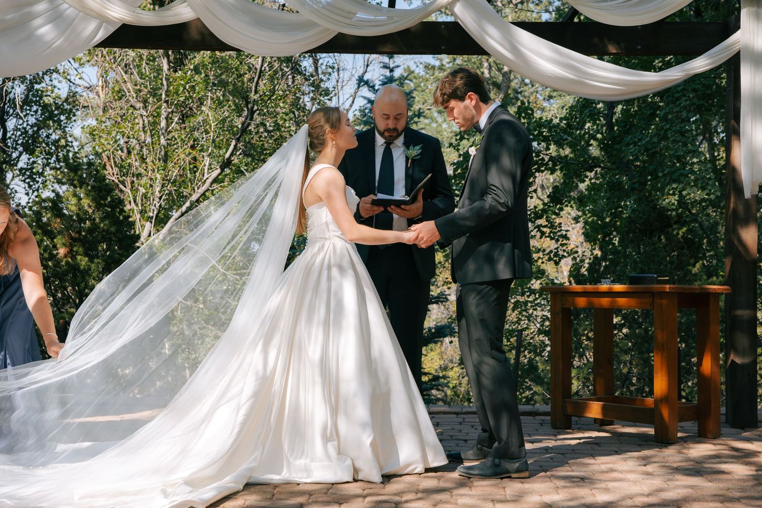 Bride and groom exchange vows outdoors under white draped canopy with officiant, surrounded by lush green trees.