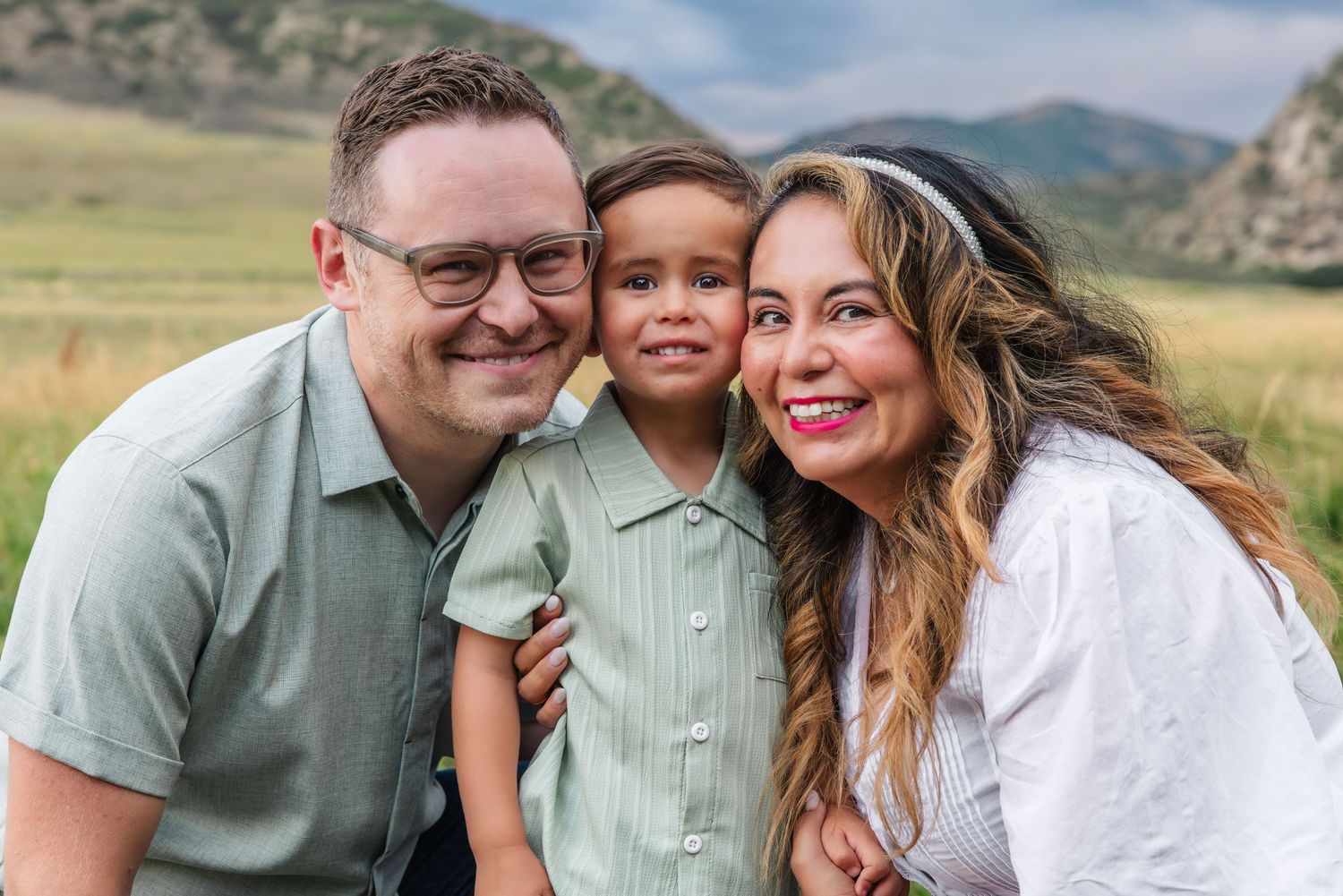 A happy family of three smiling outdoors with green hills in the background on a sunny day.