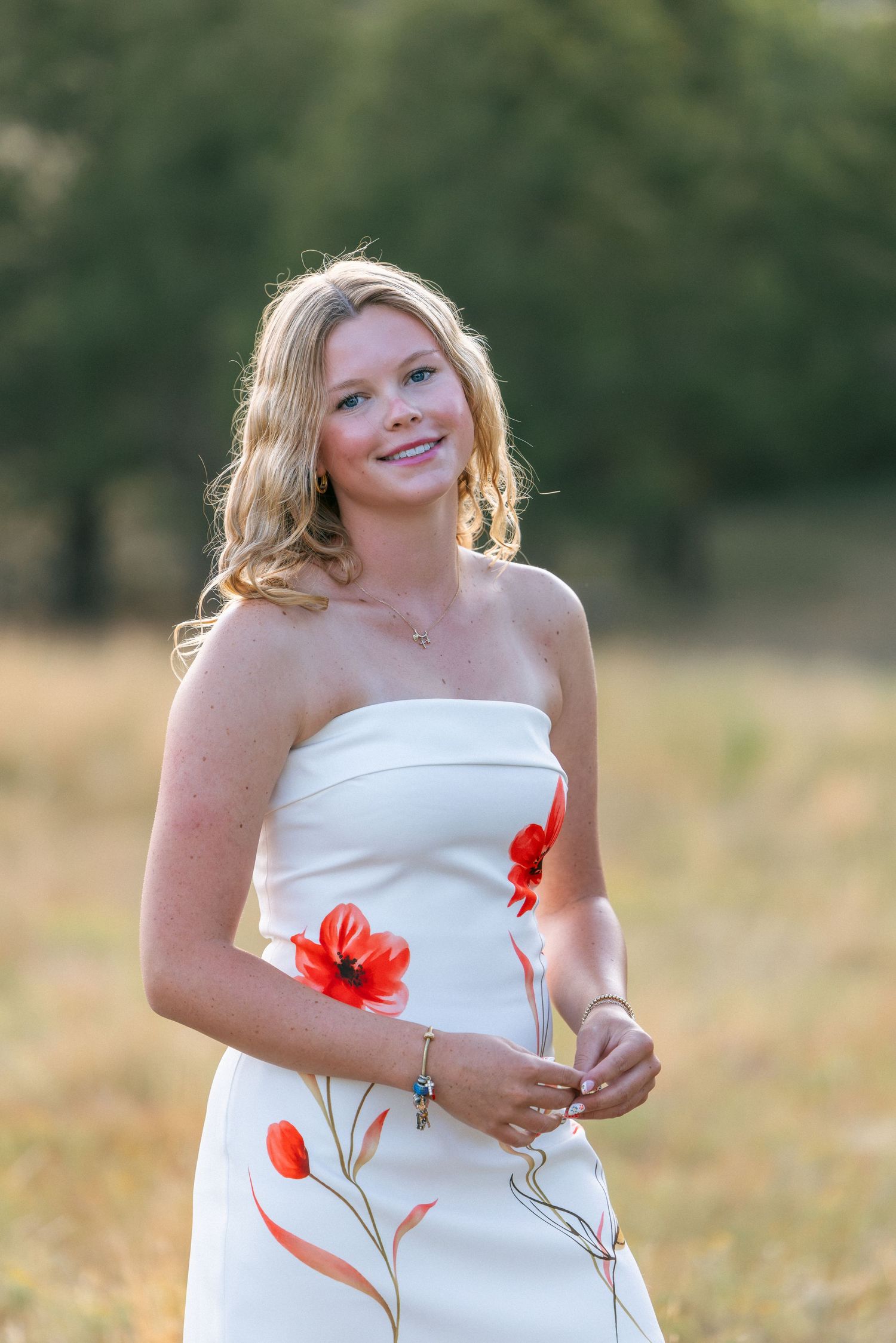 Young blonde woman smiling in white floral dress with red poppies, standing in a sunlit field.