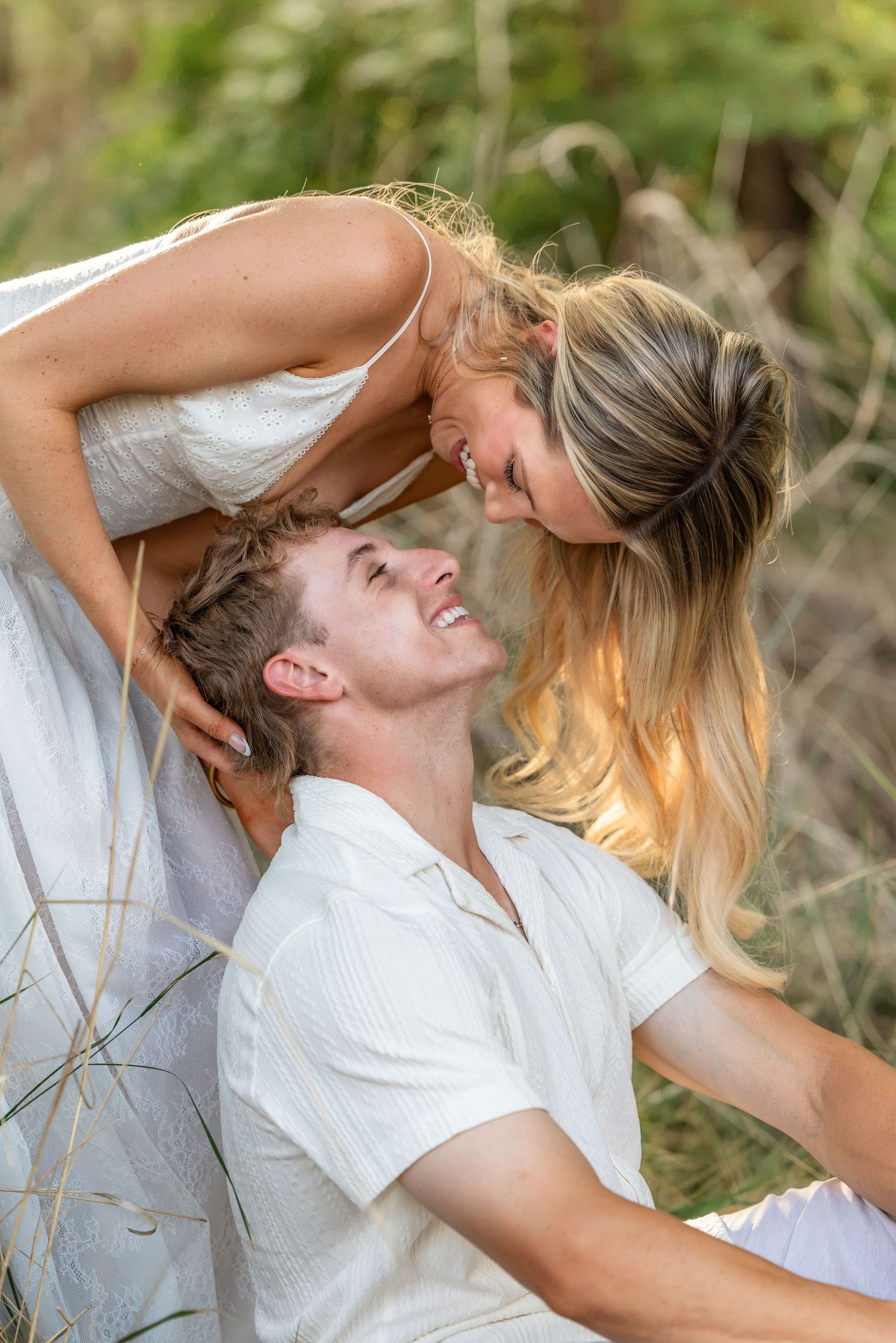 A couple in white outfits share a romantic moment outdoors, the woman leaning down to kiss the smiling man.
