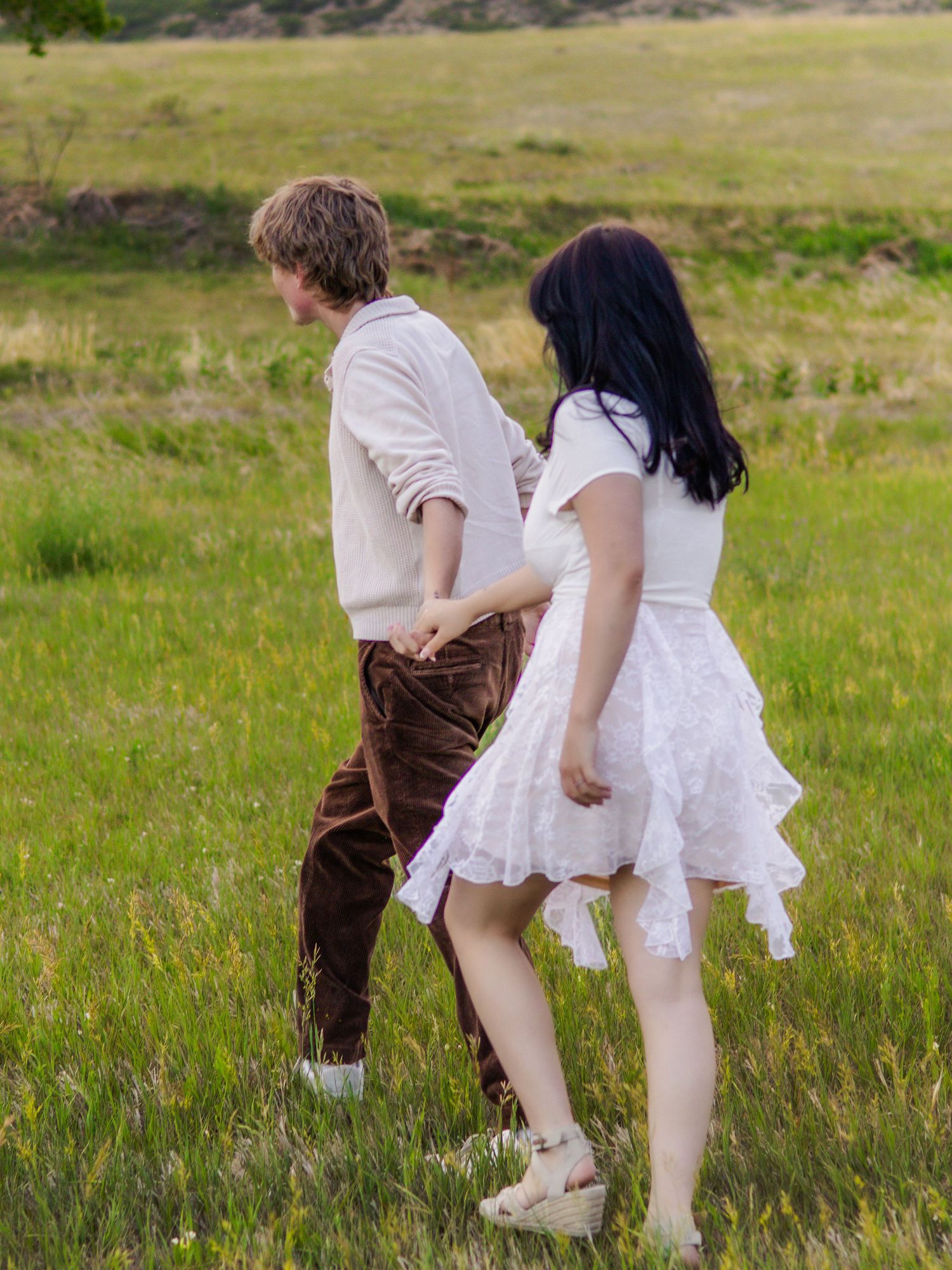 Young couple walking hand in hand through a lush green field, woman wearing white lace dress in Littleton, Colorado.