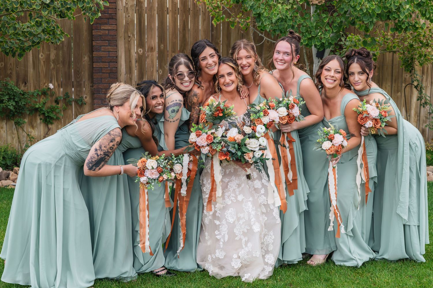 Bride in white floral gown poses with seven bridesmaids in sage green dresses holding colorful bouquets outdoors.