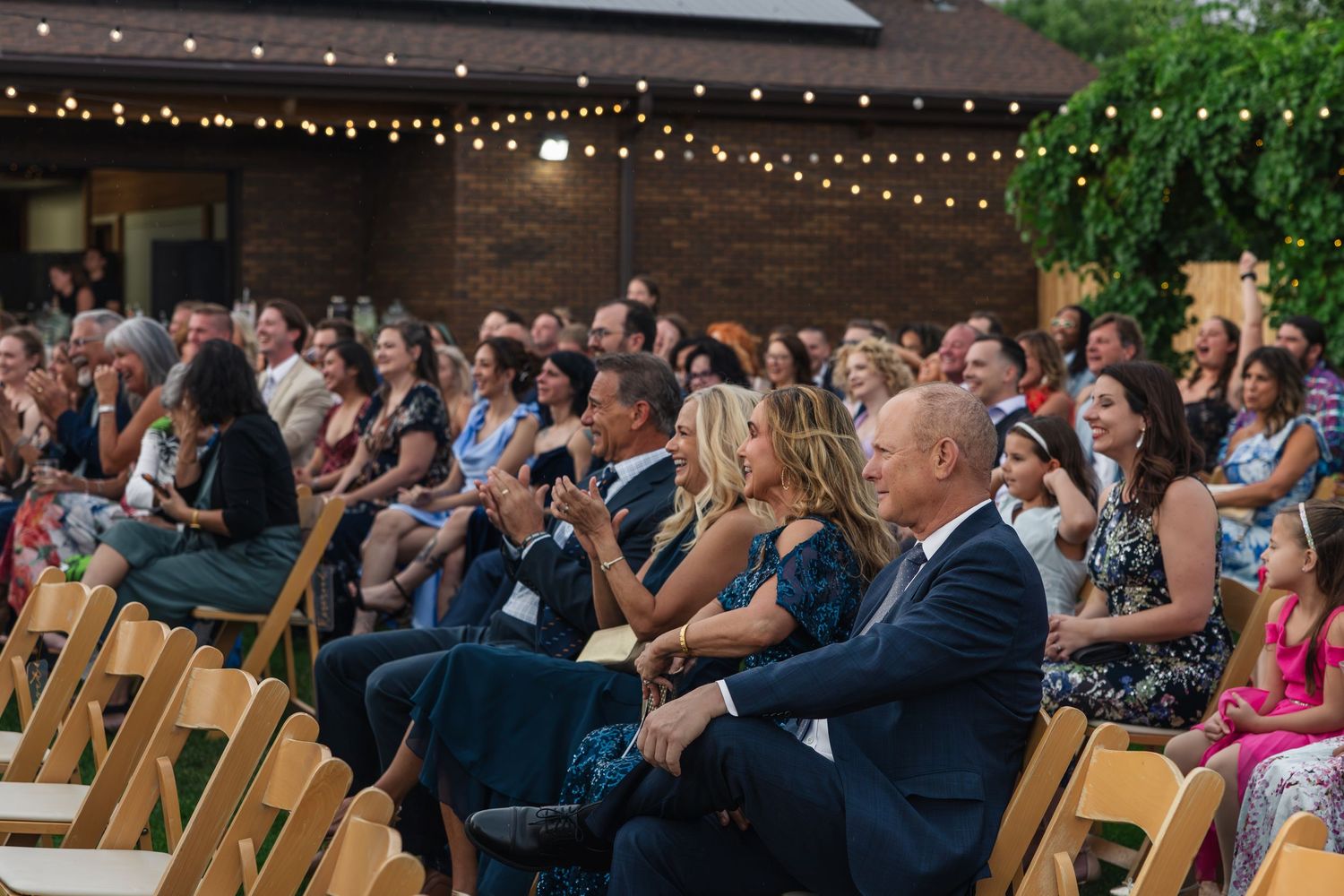 Guests seated in wooden chairs at an outdoor wedding ceremony with string lights and brick building backdrop.