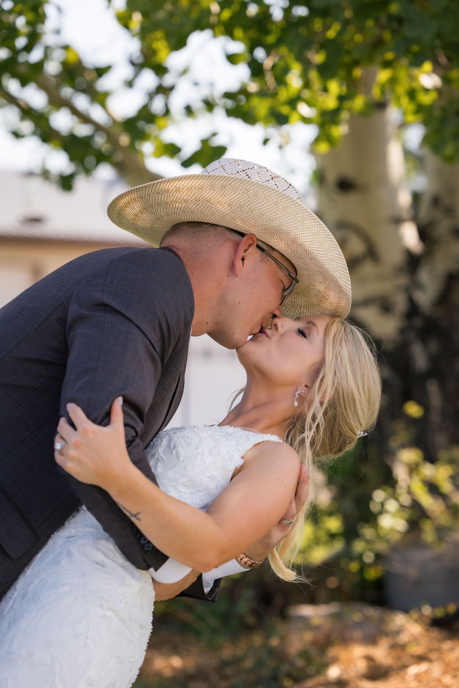 Bride and groom sharing a romantic kiss outdoors, groom wearing a cowboy hat and suit, bride in a white lace dress.