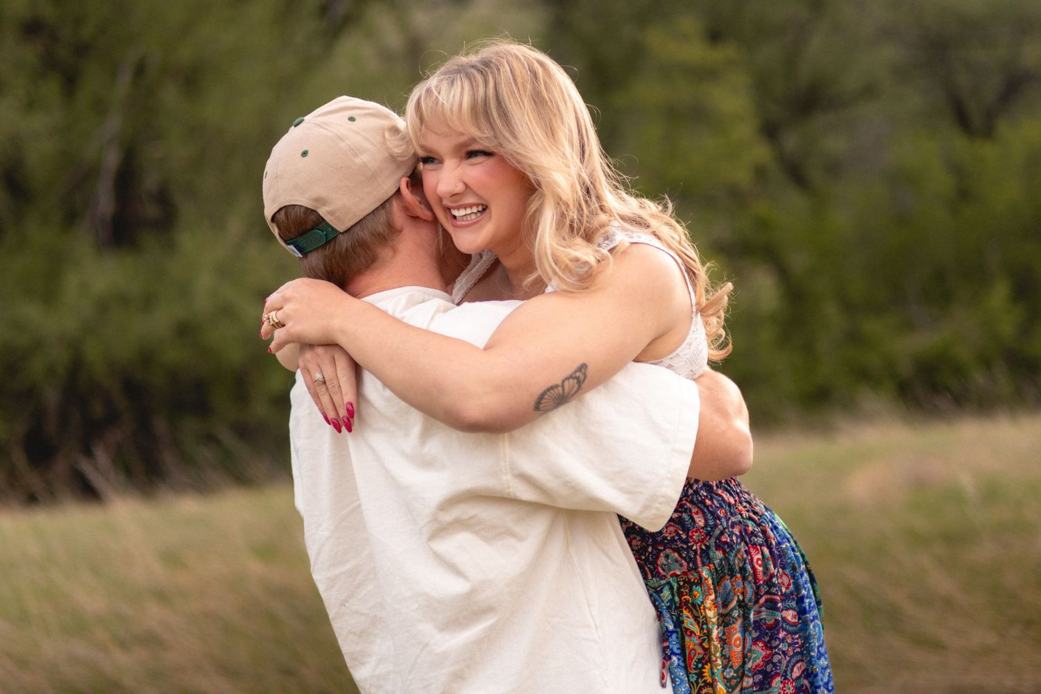 A smiling blonde woman hugs a man in a white shirt and tan cap outdoors in a green field.