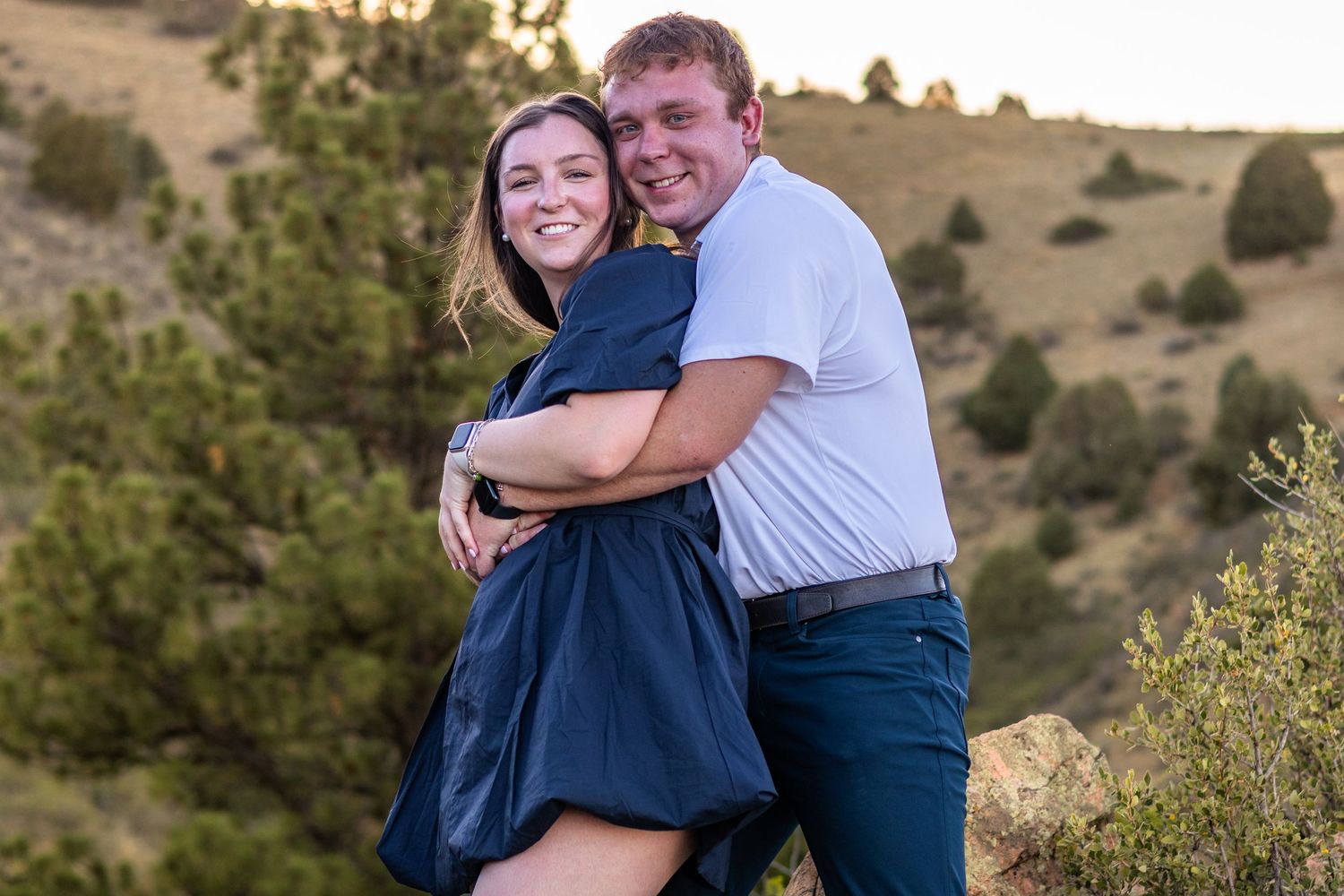 Happy couple embracing outdoors on a hillside with golden hour light and green shrubs in background.