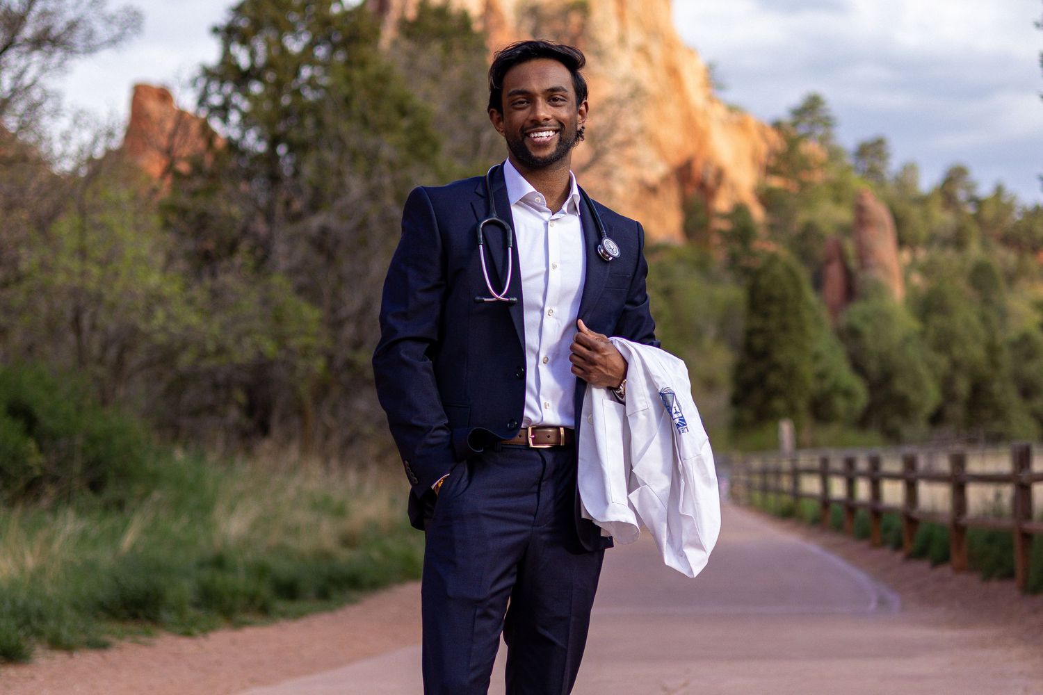 Young male doctor in navy suit holding white coat with stethoscope, standing on scenic red rock canyon path.