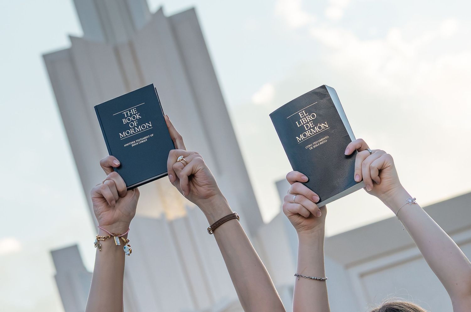 Two hands raised holding copies of The Book of Mormon in front of a white temple building.