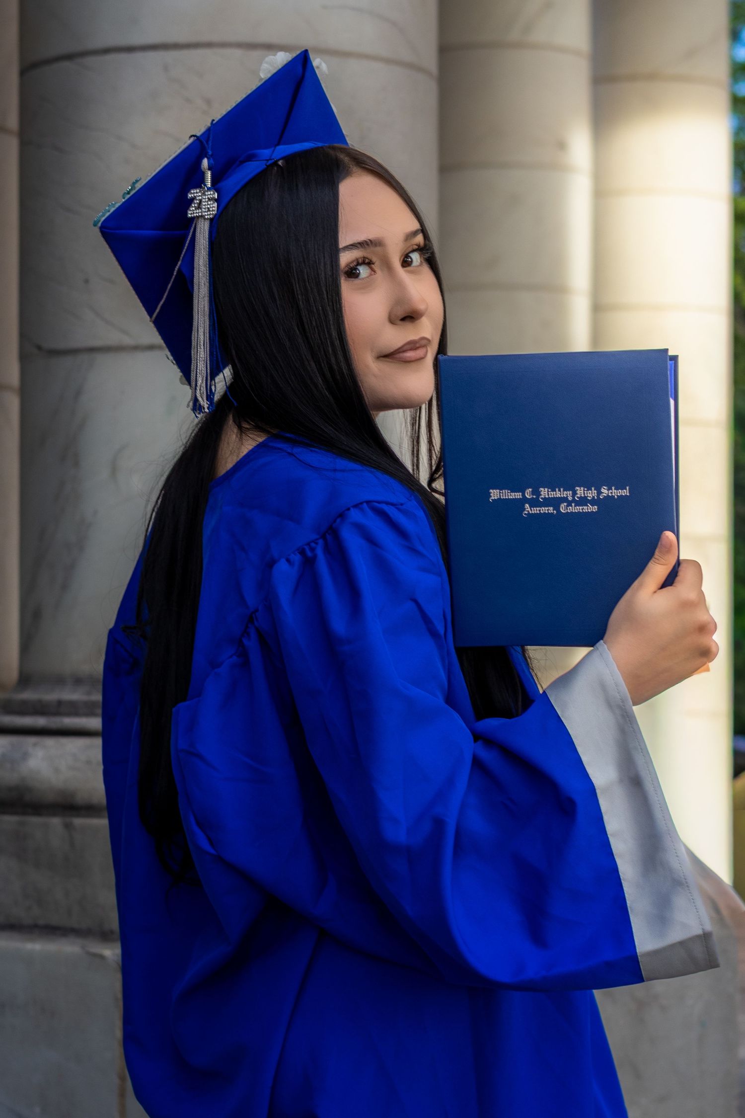 Young woman in blue graduation cap and gown holding diploma from William C. Hinkley High School, Aurora, Colorado.