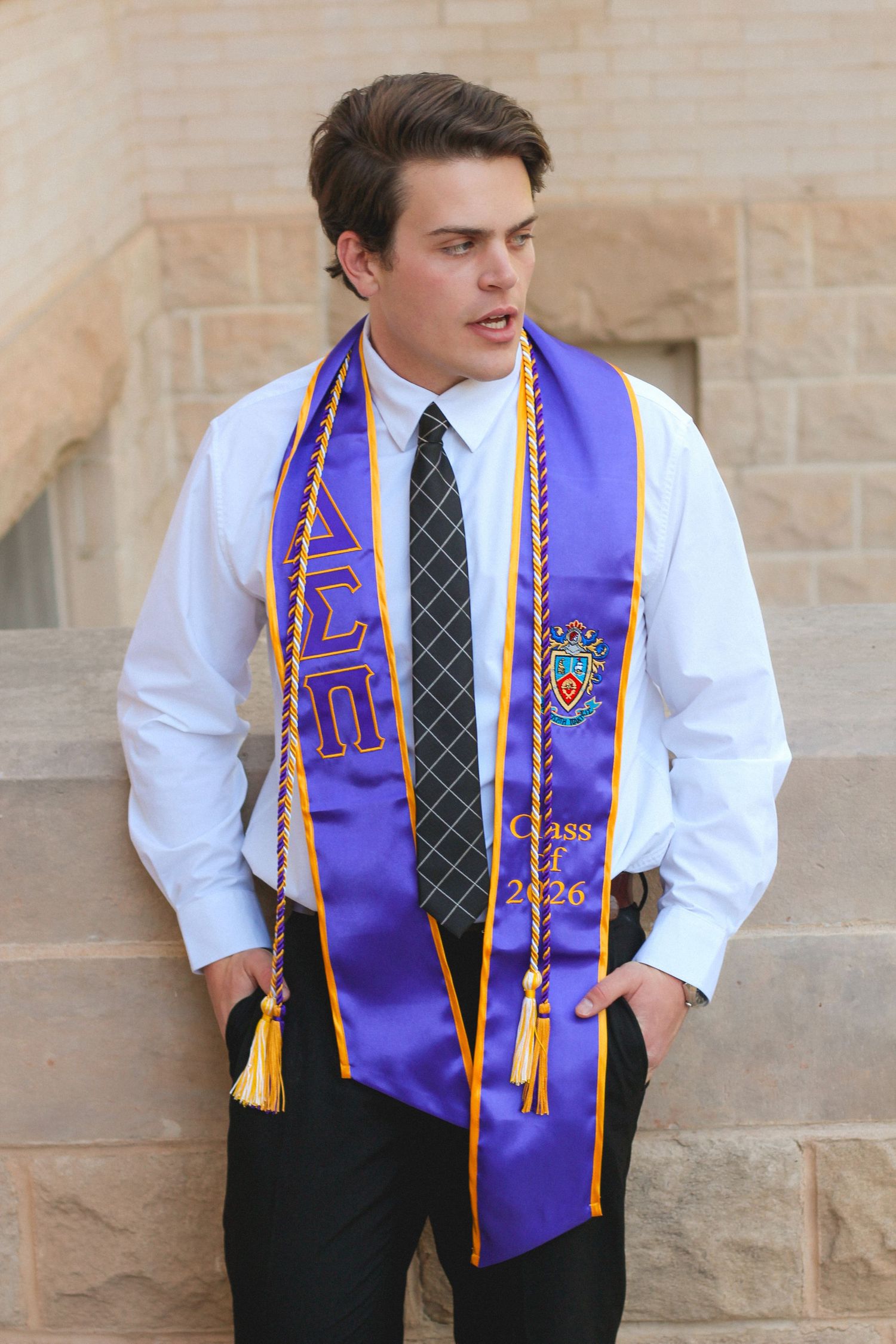 Young man in purple and gold Class of 2026 graduation stole leans against stone building wall.
