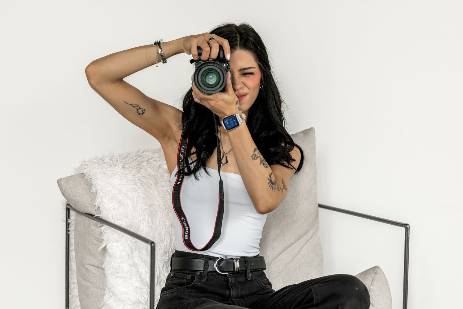 Young woman with tattoos sits on a couch, holding a DSLR camera up to her face, wearing a white tank top.