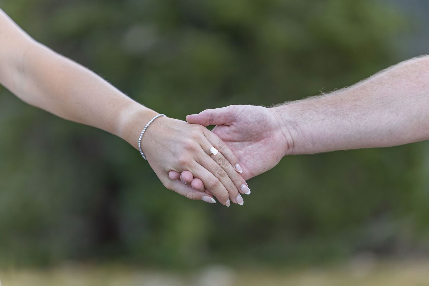Two people gently holding hands outdoors, one wearing a gold bracelet, with a blurred green background.