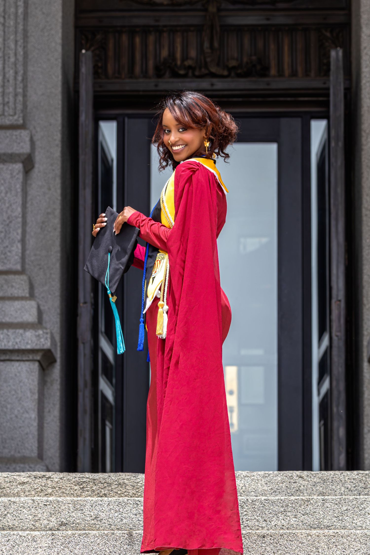 Smiling graduate in red doctoral gown with colorful stoles stands at ornate building entrance in Littleton, Colorado.