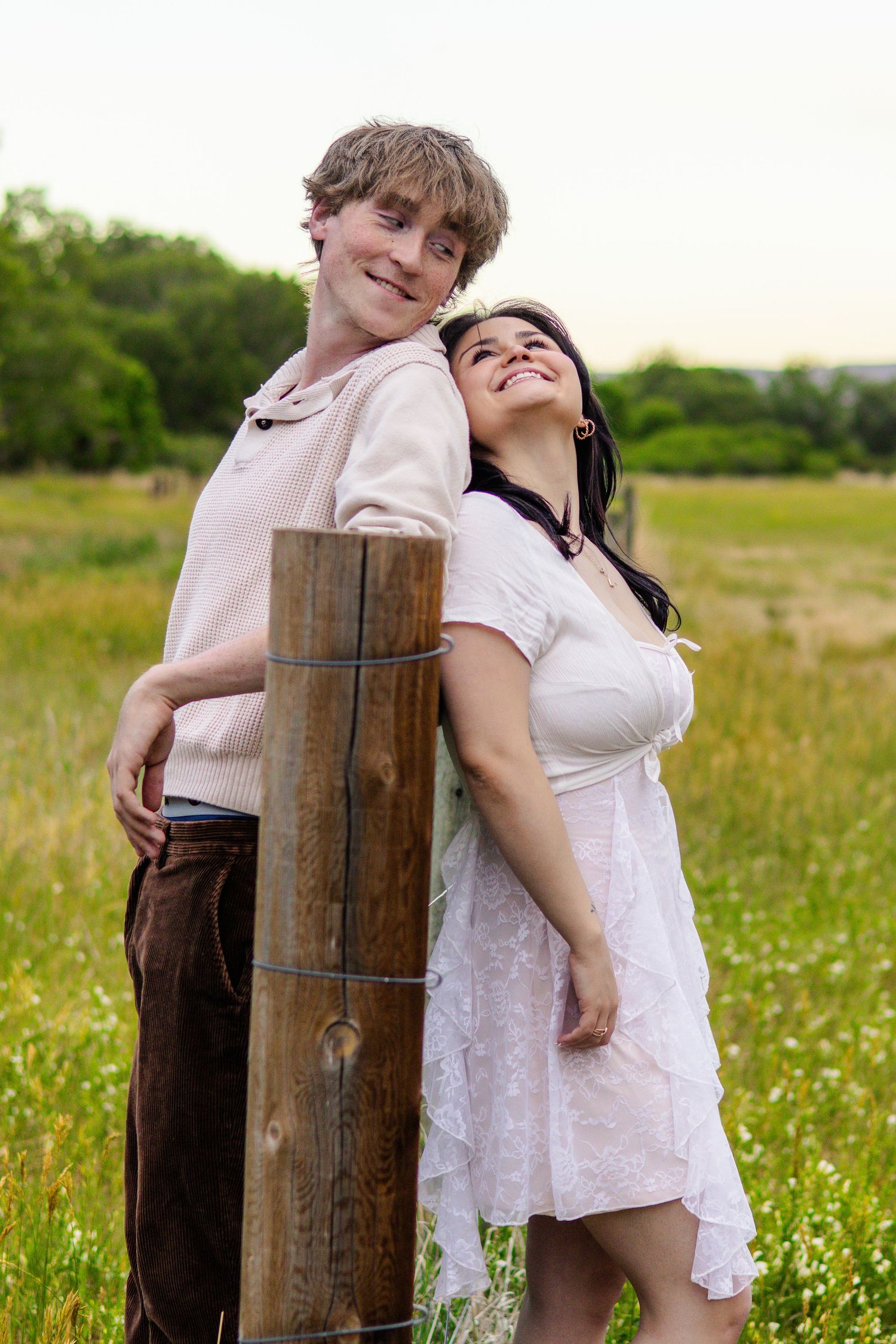 A happy young couple poses back-to-back against a wooden fence post in a scenic green countryside field in Littleton, Colorado.