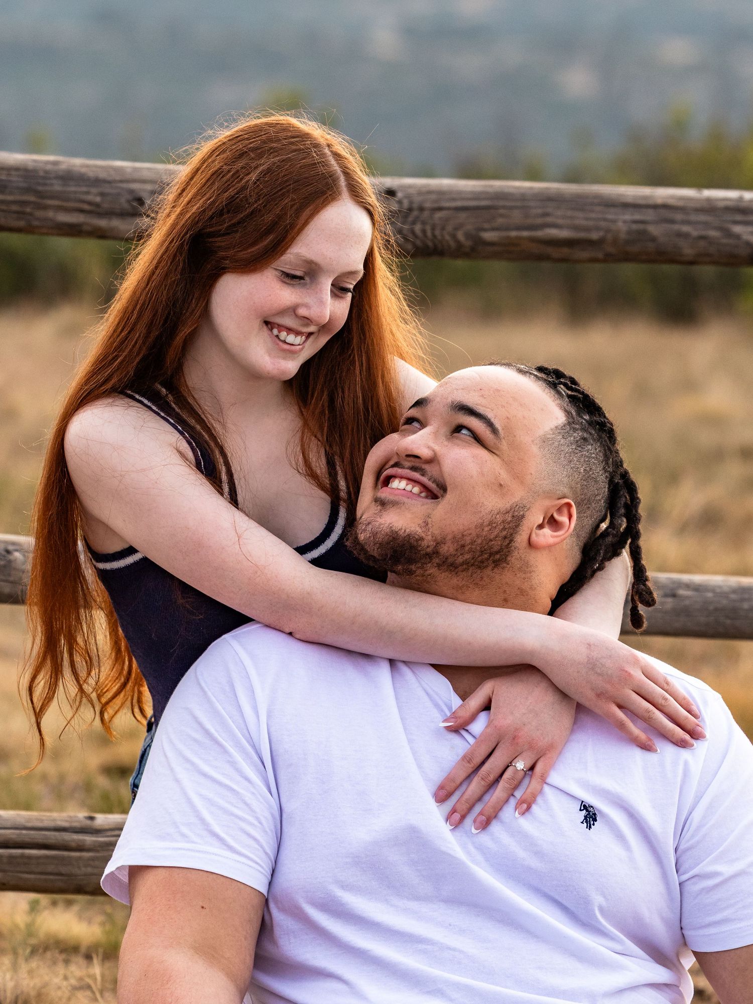 A smiling redhead woman embraces a man with braids from behind near a rustic wooden fence in an outdoor field.