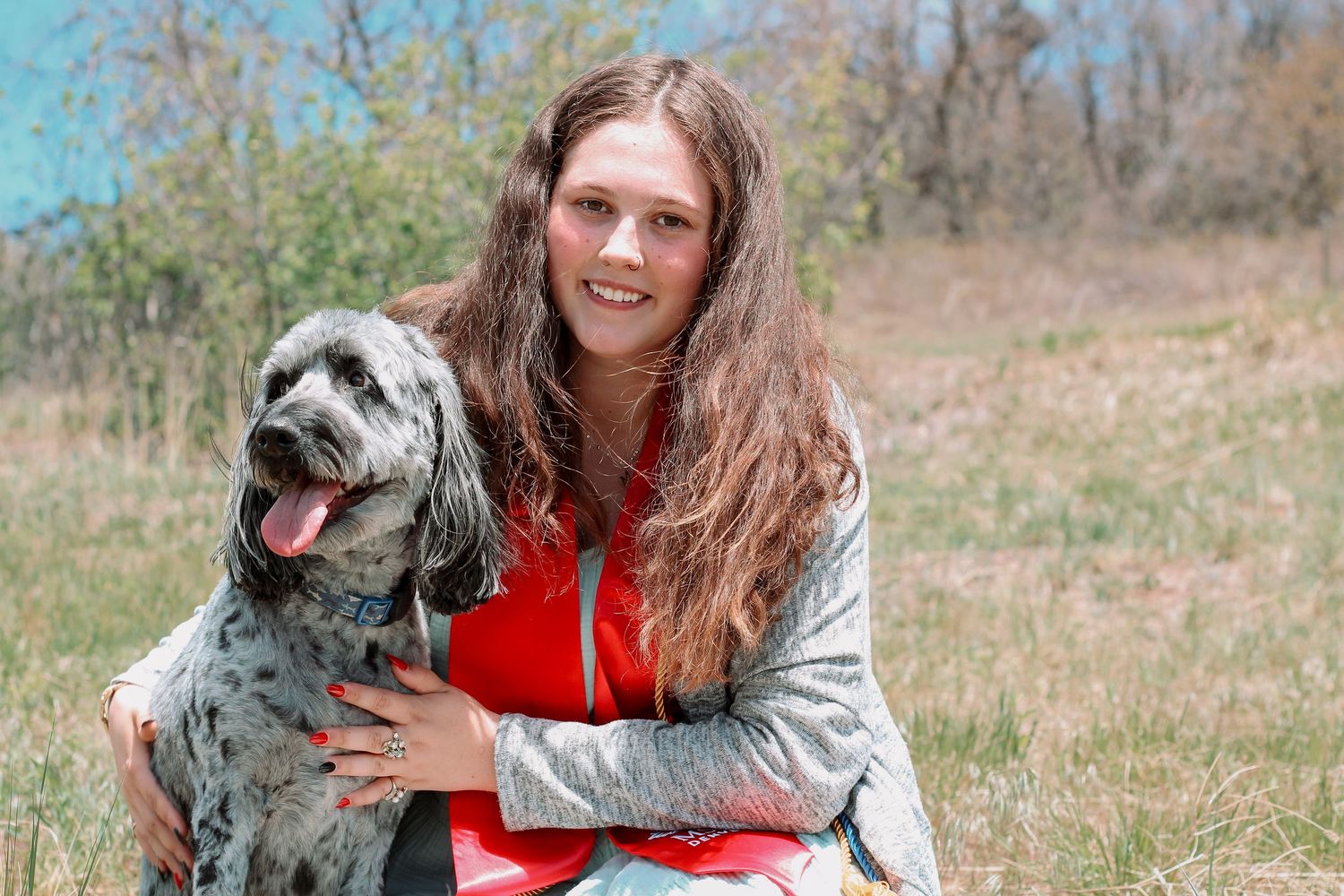 Young woman sitting in grassy field with her gray spotted dog on sunny day.