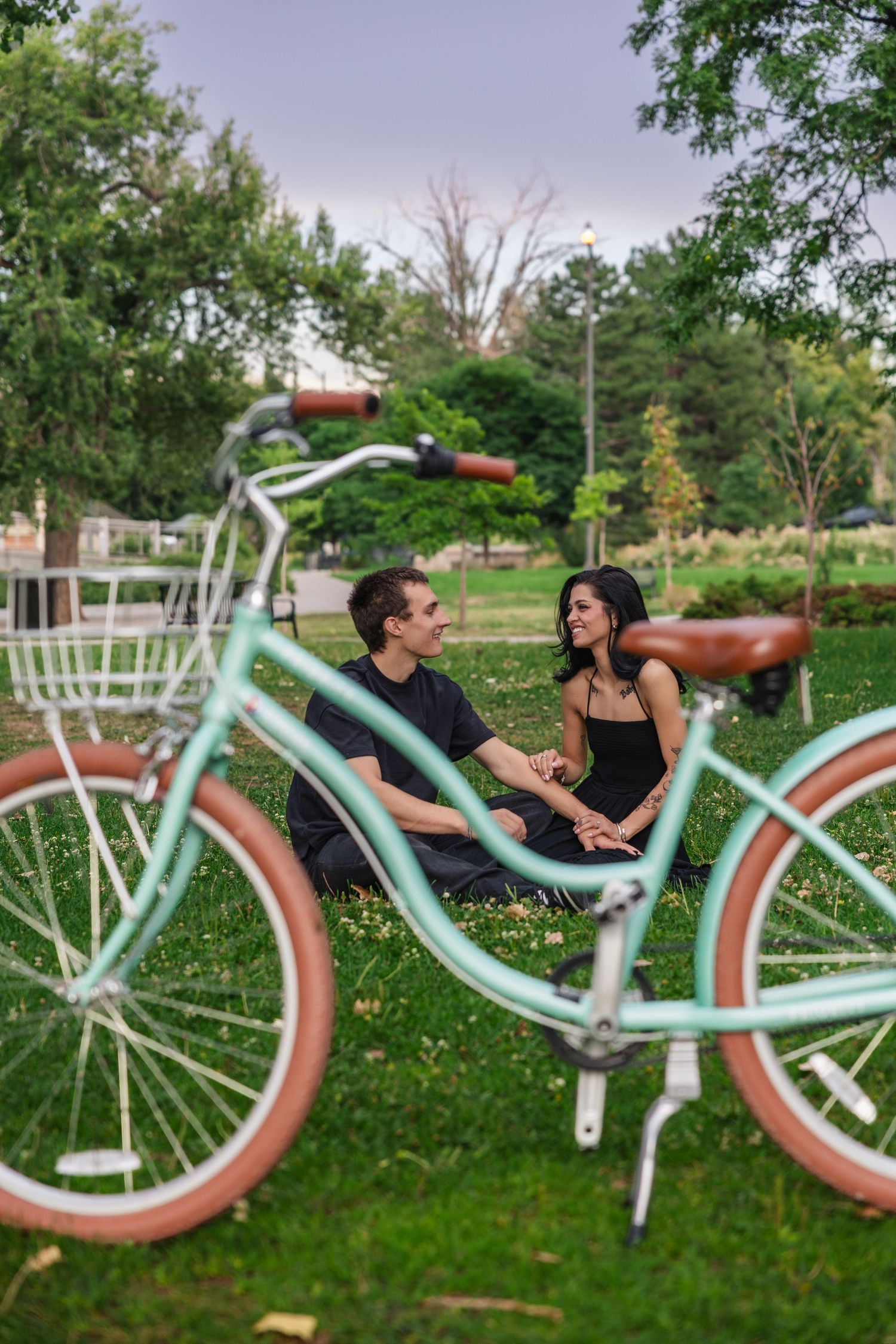 Couple sitting on green grass in a park holding hands beside a mint green cruiser bicycle on a cloudy day.