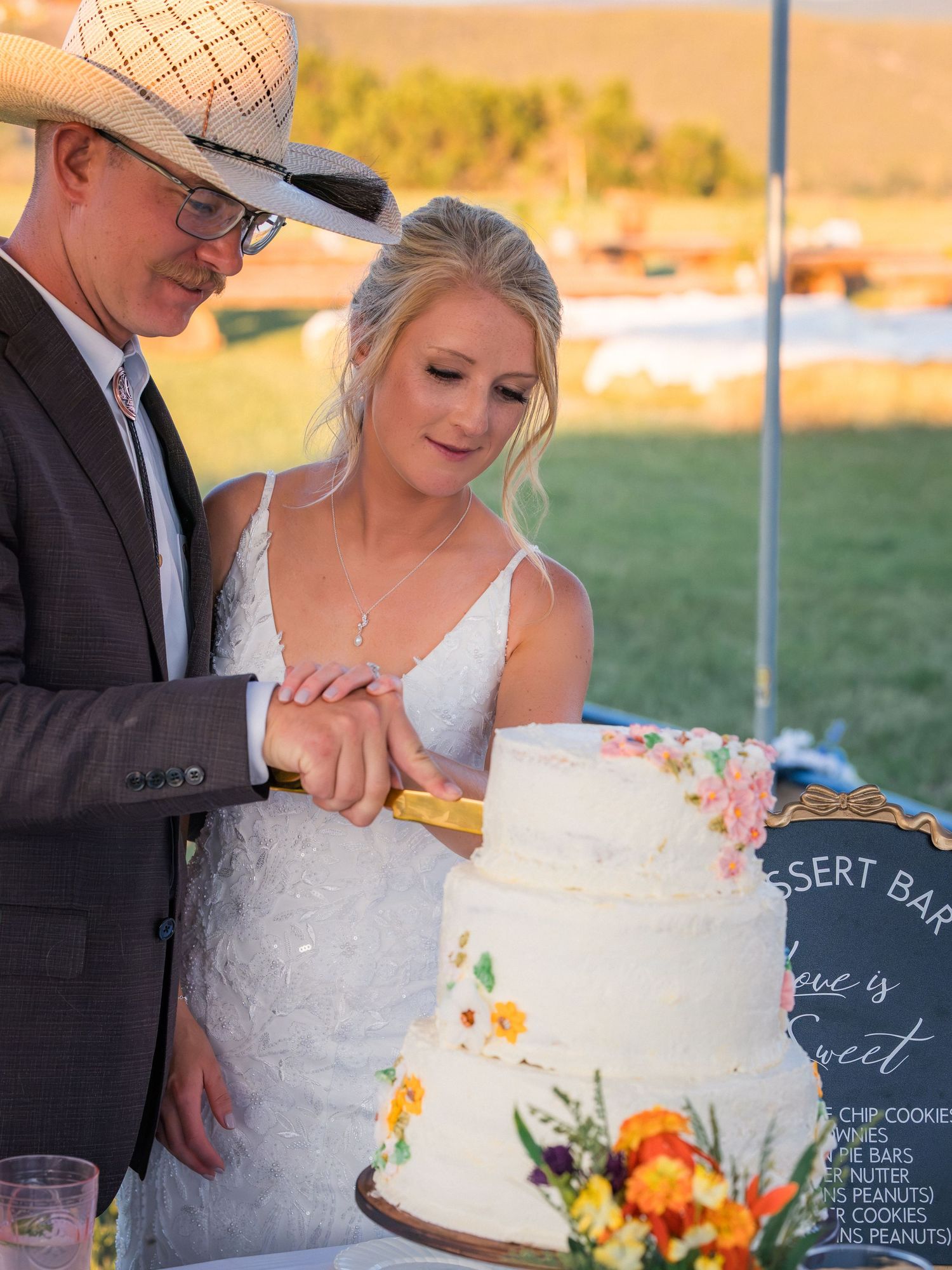 Bride and groom cutting a white floral wedding cake outdoors at a rustic country wedding reception.