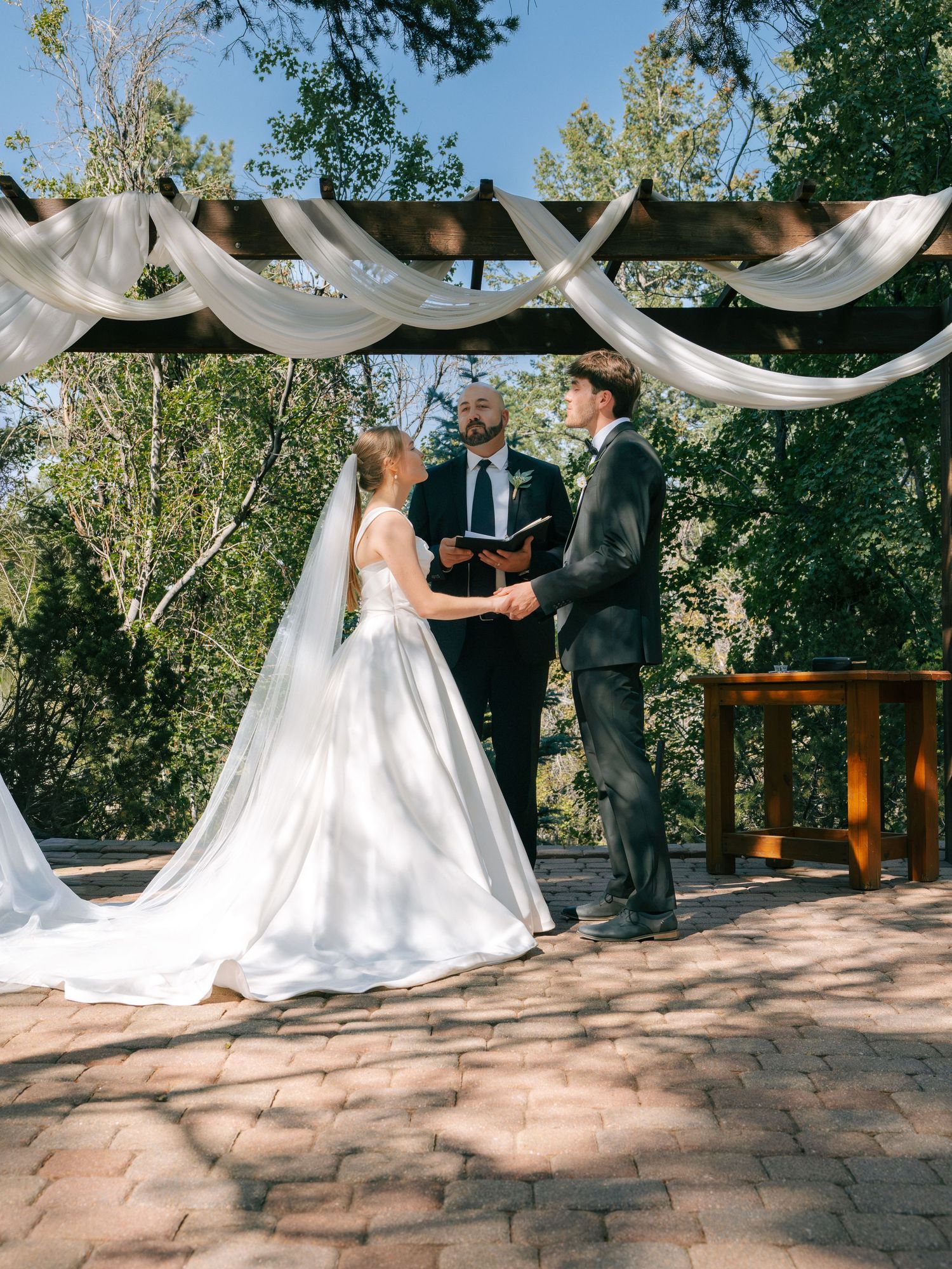 Outdoor wedding ceremony with bride in white gown and groom in gray suit exchanging vows under draped pergola.