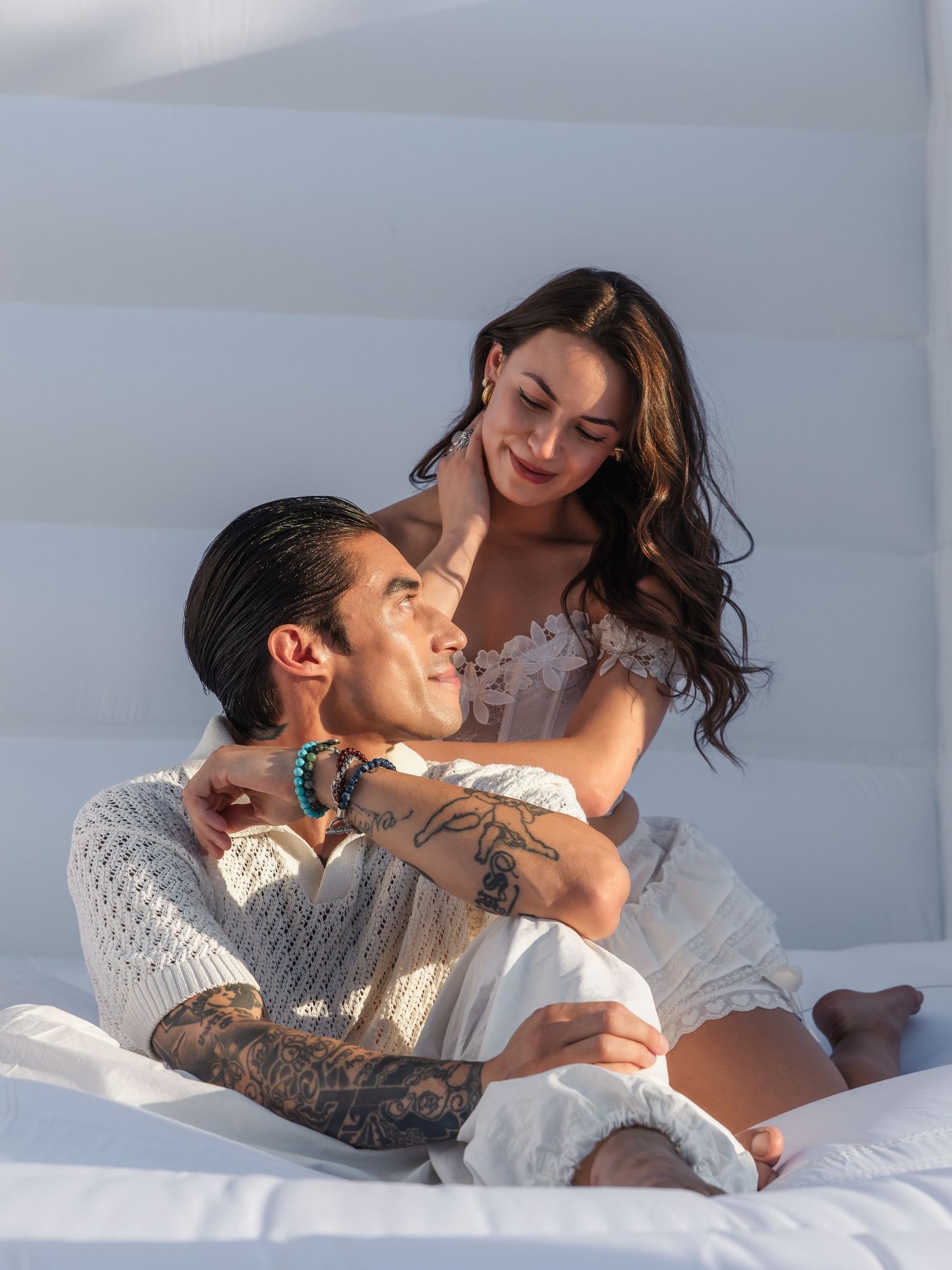 A tattooed man in white sits on a bed while a brunette woman in a white dress embraces him from behind.
