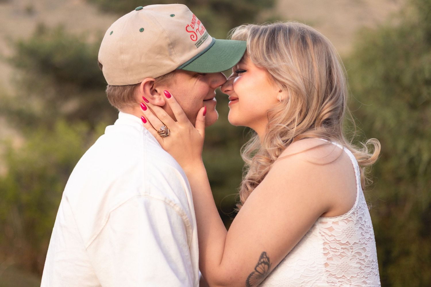 Couple sharing a romantic kiss outdoors, woman with blonde hair and tattoo, man in green cap.