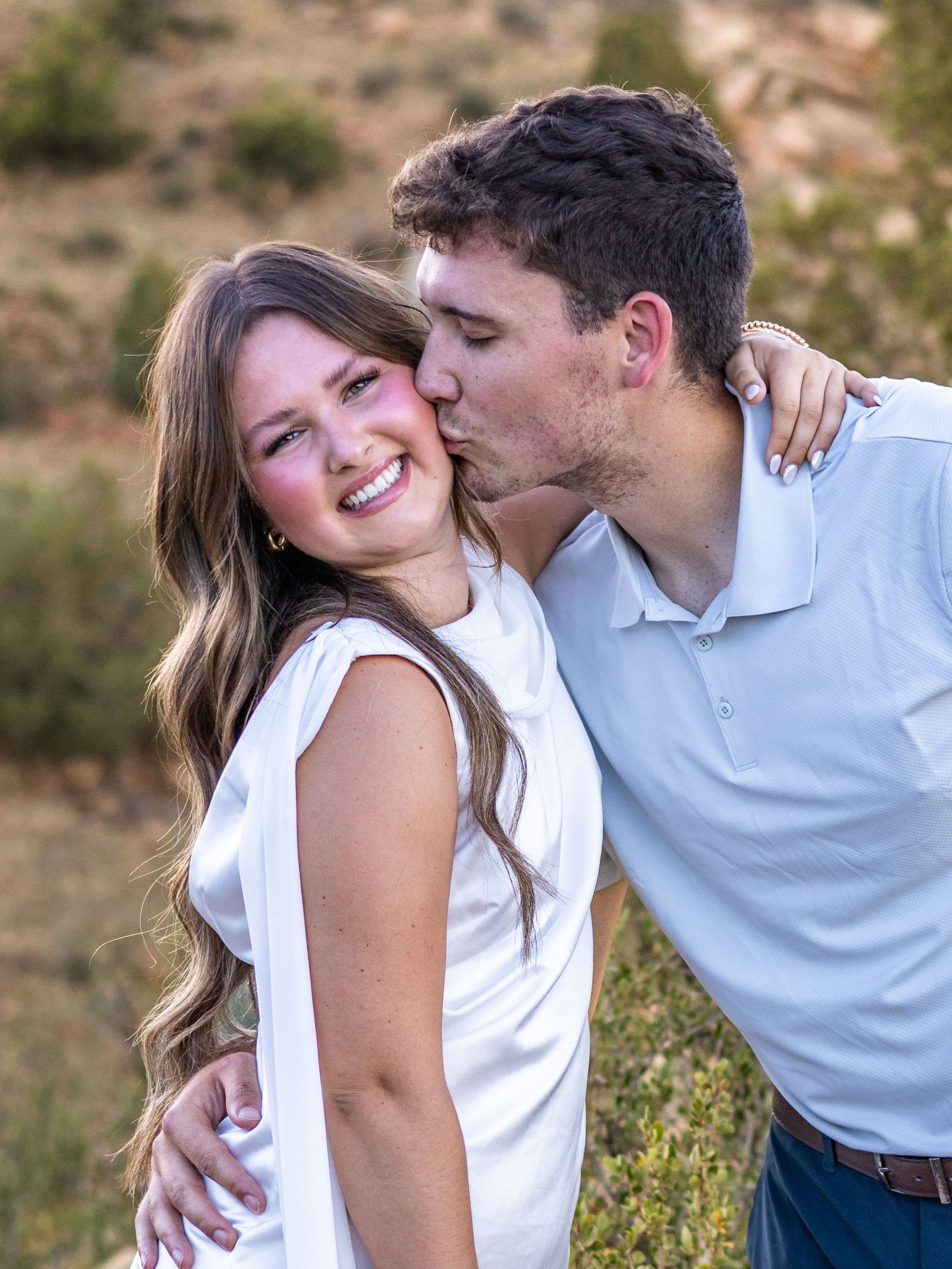 A young couple poses outdoors, woman smiling in white dress while man kisses her cheek affectionately.