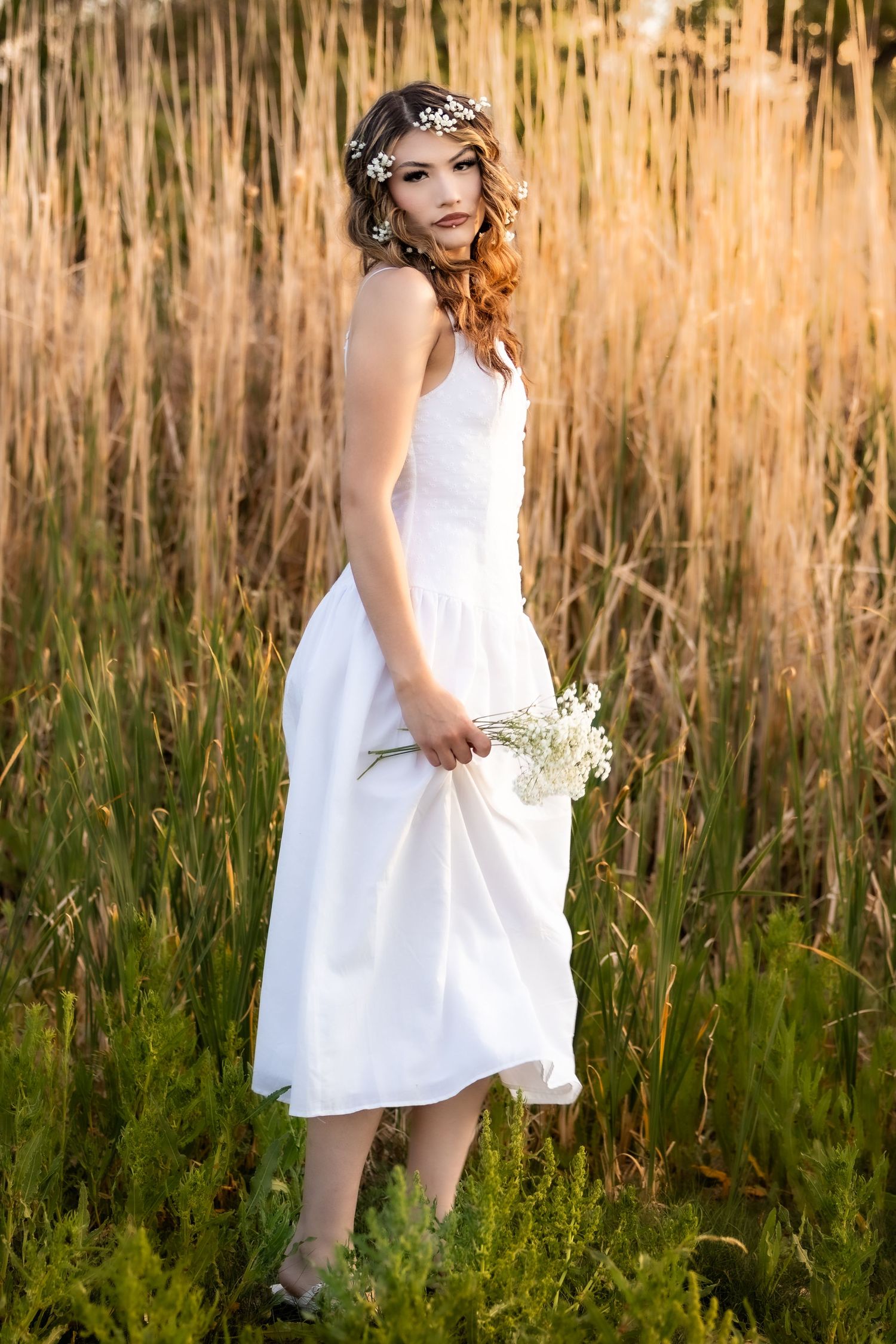 Young woman in white midi dress and floral crown holds wildflowers while standing in golden sunlit tall grass field.