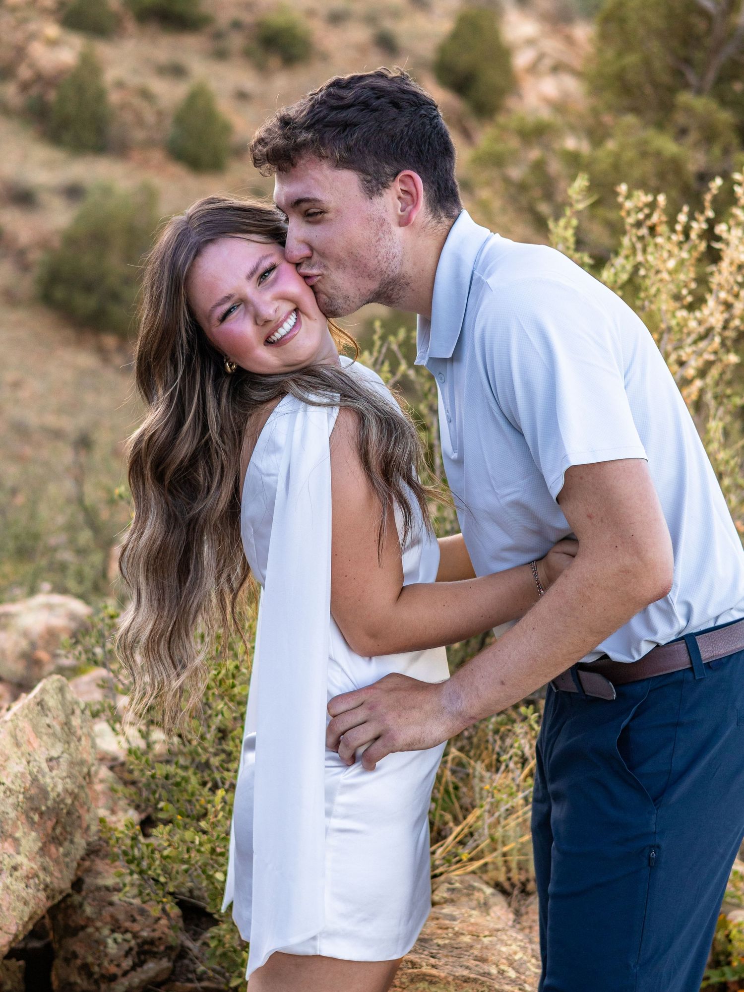 Couple in white outfits sharing a romantic kiss outdoors among rocks and greenery in a scenic mountain setting.