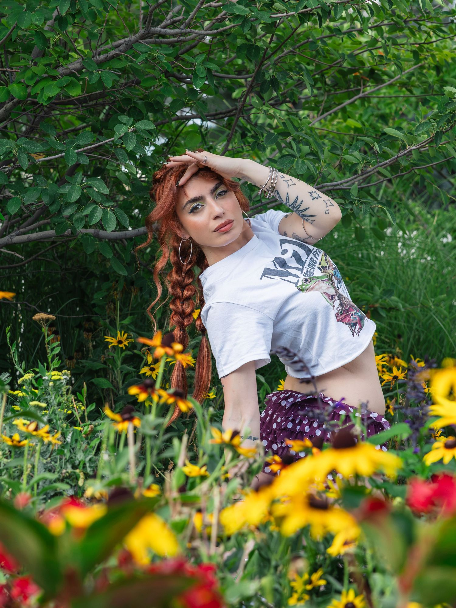 Red-haired woman with braids and tattoos posing among yellow wildflowers in a lush green garden.