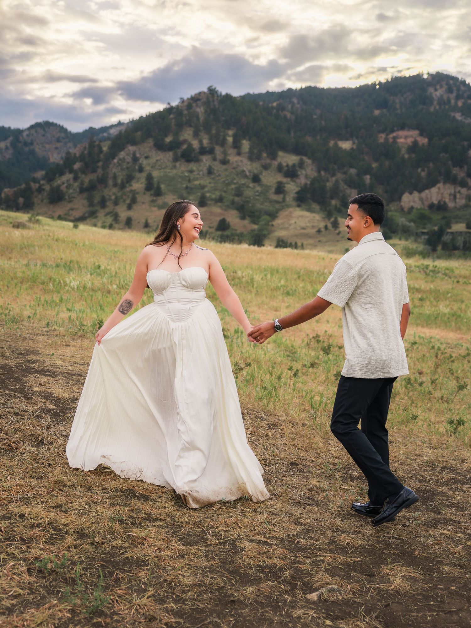 A joyful couple holds hands in a mountain meadow, the bride in a flowing white gown, groom in white shirt and black pants.