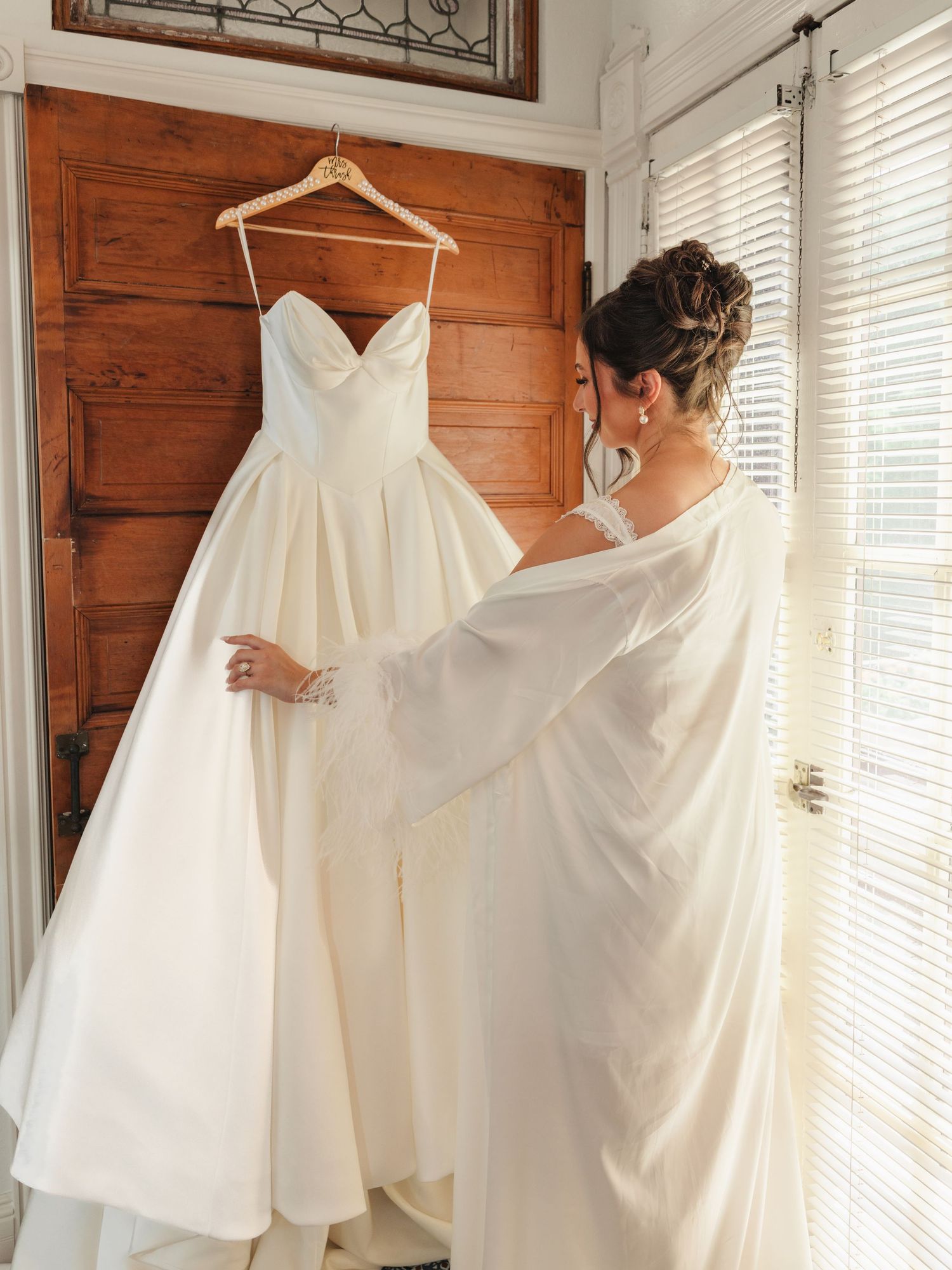 Bride in white robe admiring elegant strapless ball gown wedding dress hanging on wooden door.