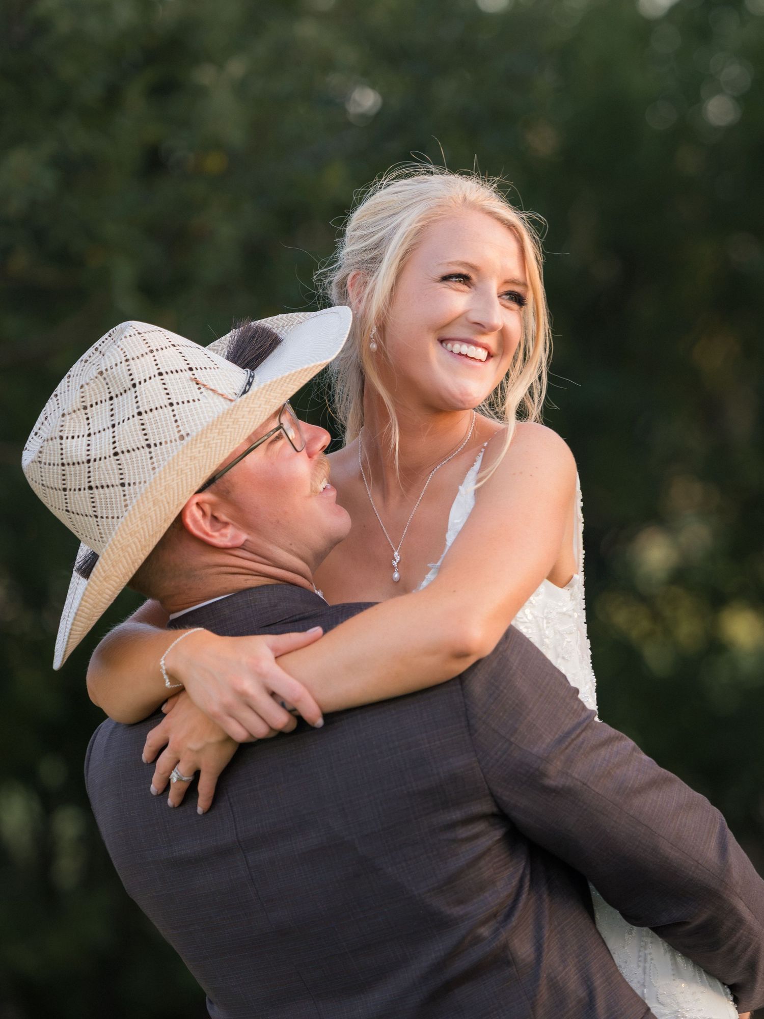 A groom in a straw hat lifts his smiling blonde bride outdoors during a sunny wedding celebration.
