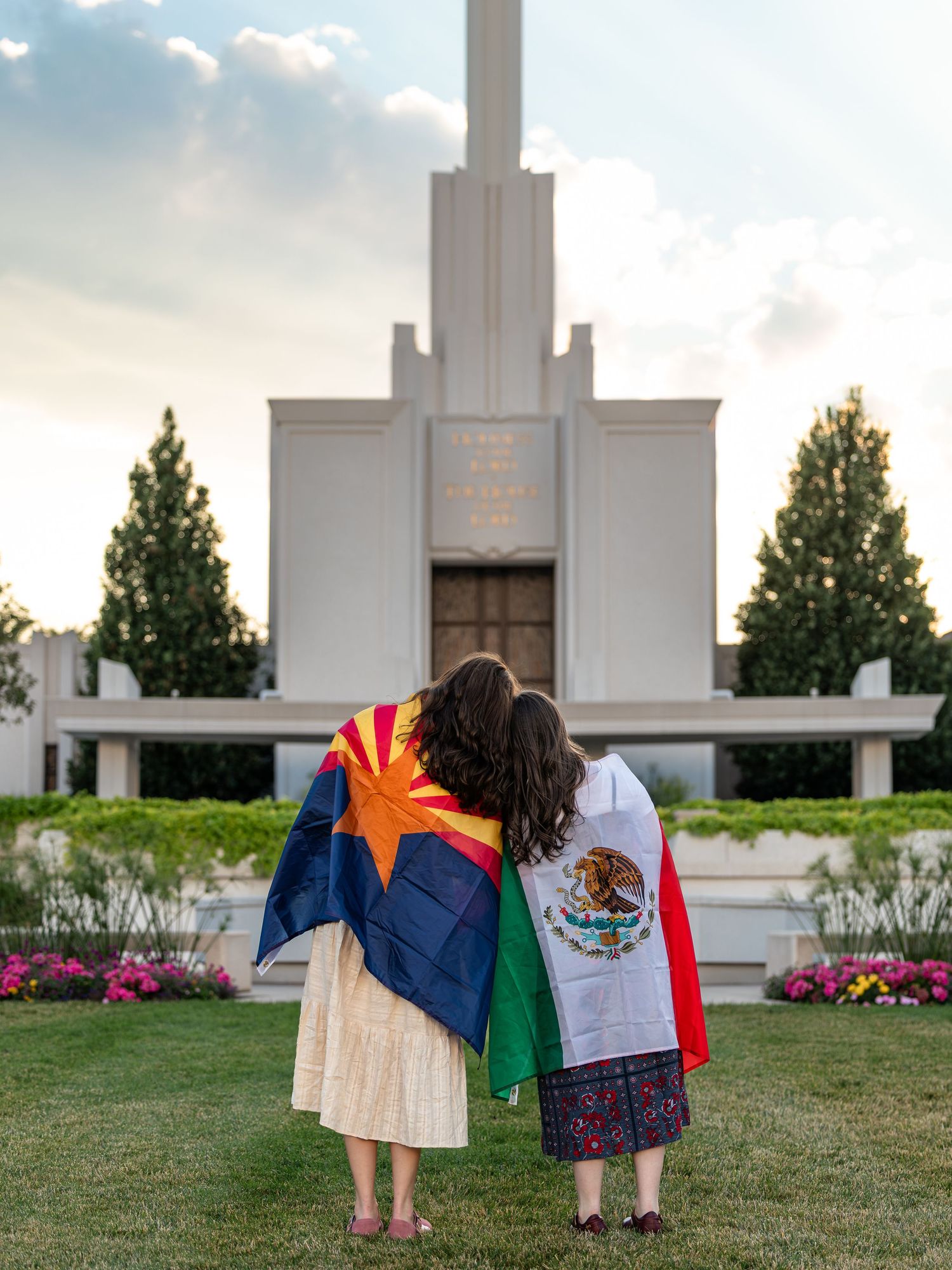 Two women draped in Arizona and Mexican flags stand before an LDS temple at sunset.