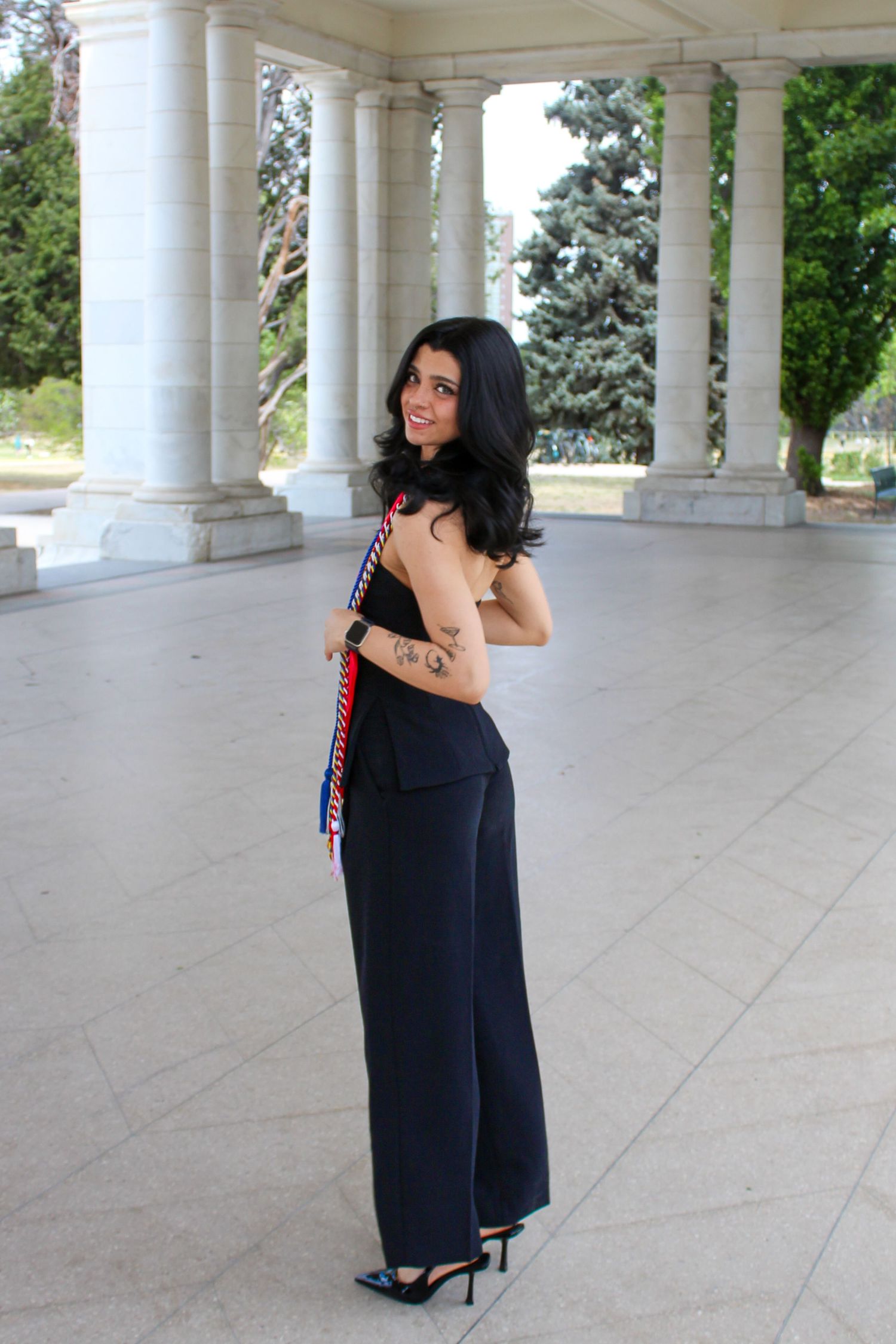 Graduate poses sideways showing tattoo and colorful honor cords at an elegant columned outdoor venue.