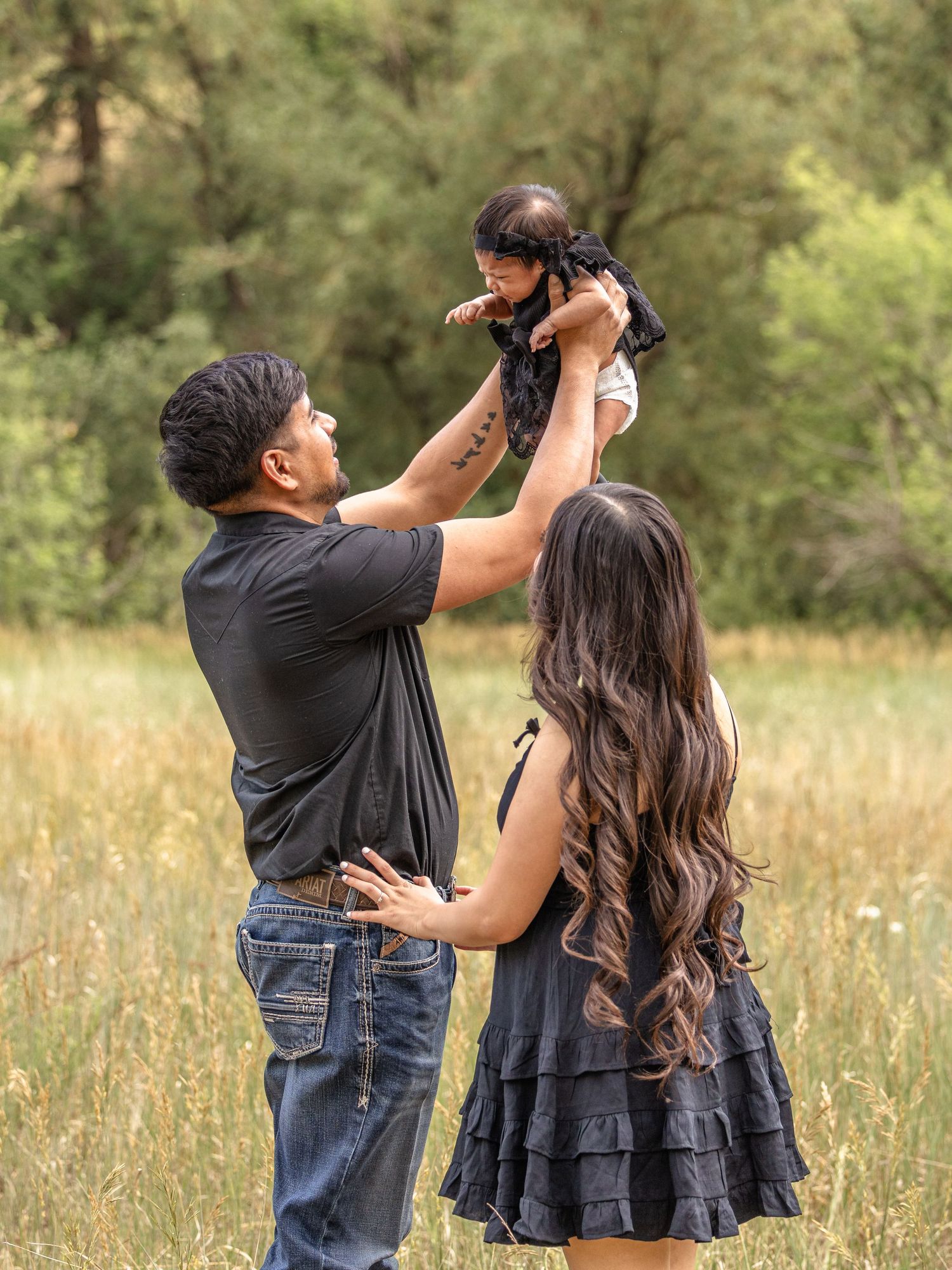 A couple in matching black outfits lifts their baby girl in a sunlit meadow during a family photo session.