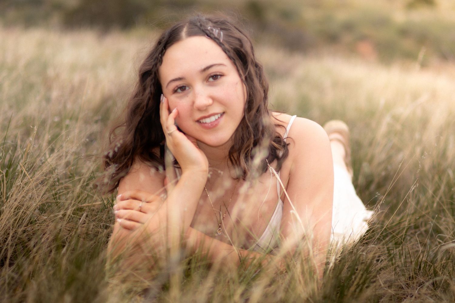 Smiling young woman with curly brown hair lying in tall golden grass, resting on hand in Littleton, Colorado.