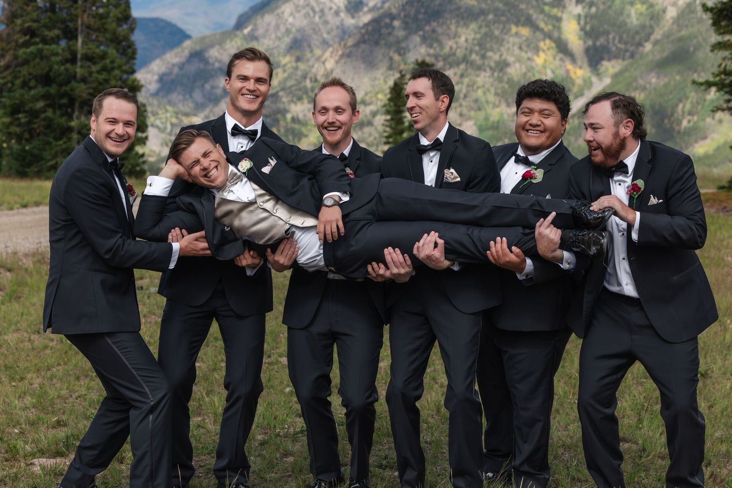 Groomsmen in black tuxedos playfully lifting one man outdoors with mountain and pine tree backdrop at a wedding.