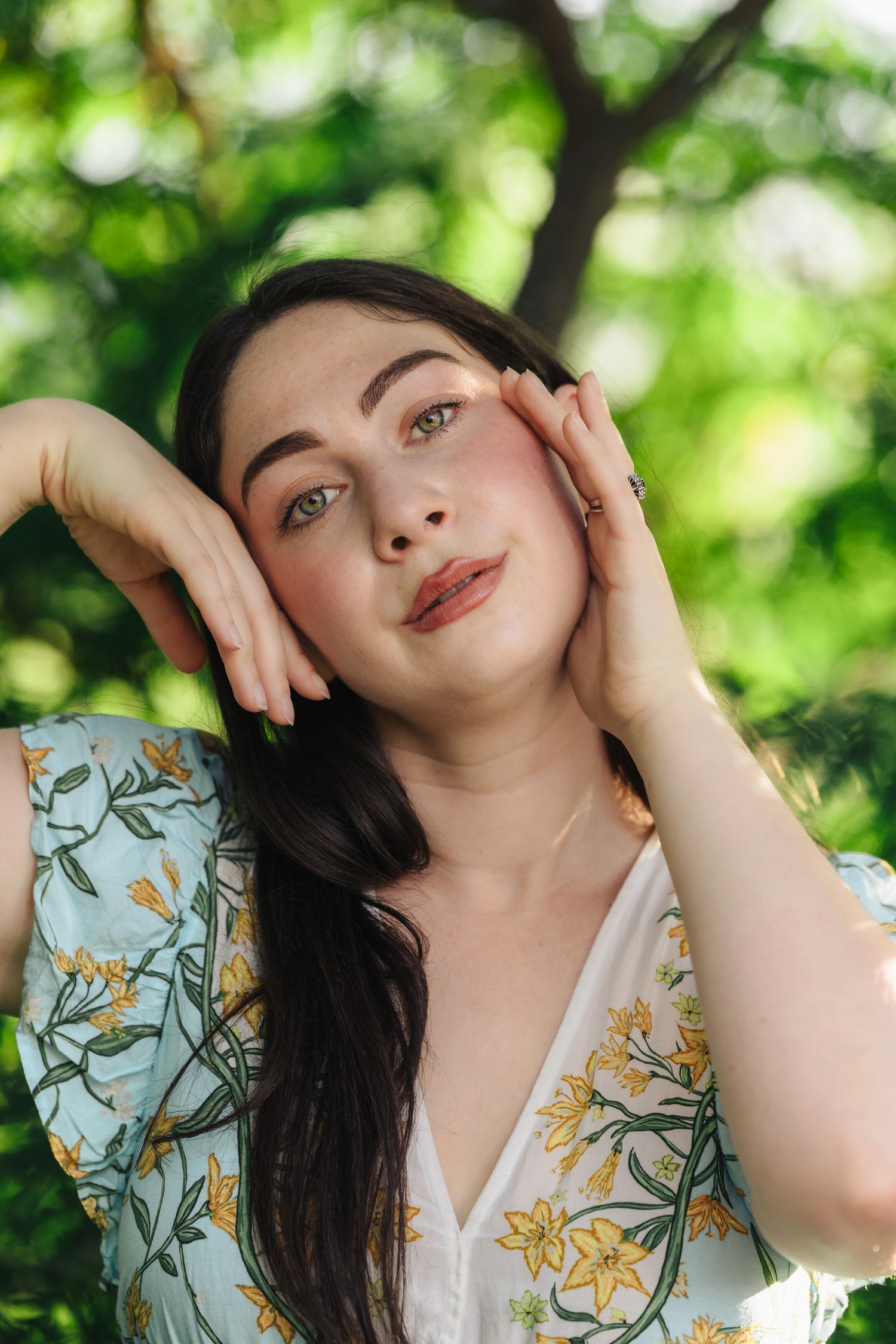 Young woman with dark hair in floral dress poses outdoors among green trees, hands framing her face.