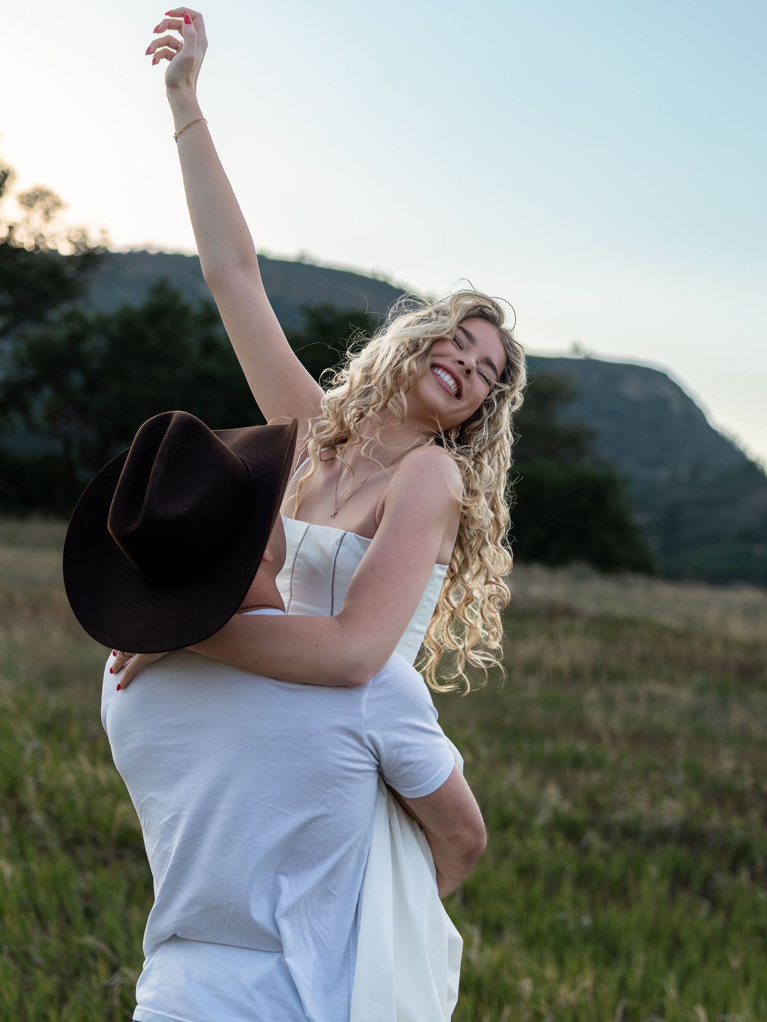 A joyful blonde woman in a white dress raises her arm while being lifted by a partner in a scenic field.
