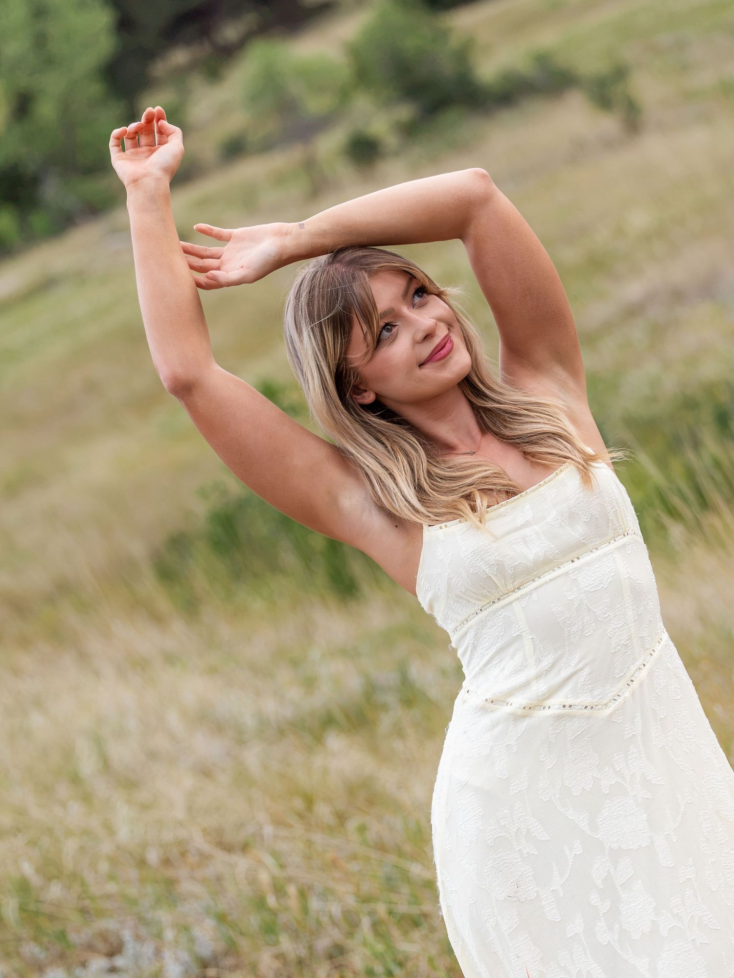 A woman in a white dress poses gracefully outdoors with arms raised, smiling in a sunlit grassy field.