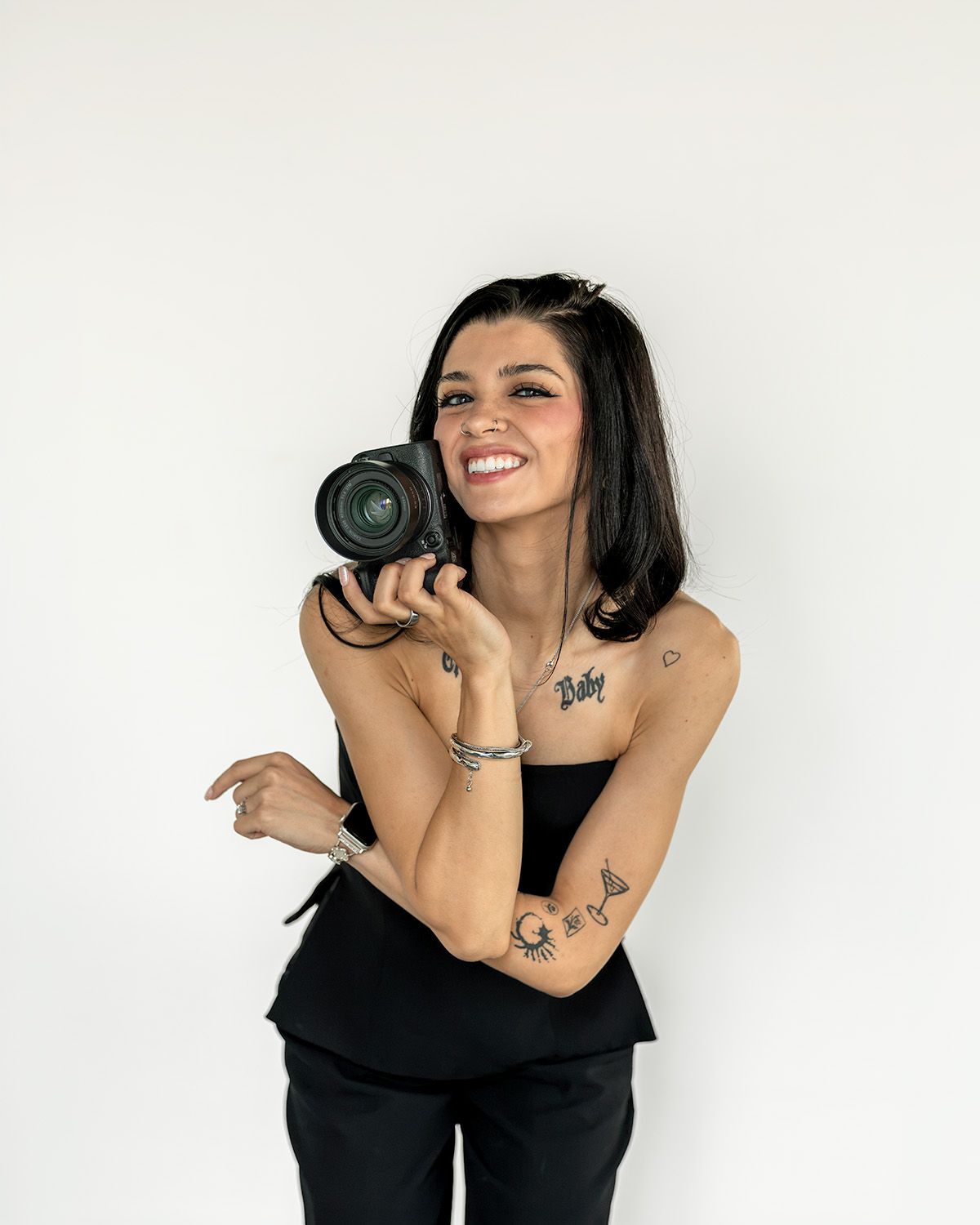 Smiling woman with tattoos holding a DSLR camera, wearing a black top against a white background.