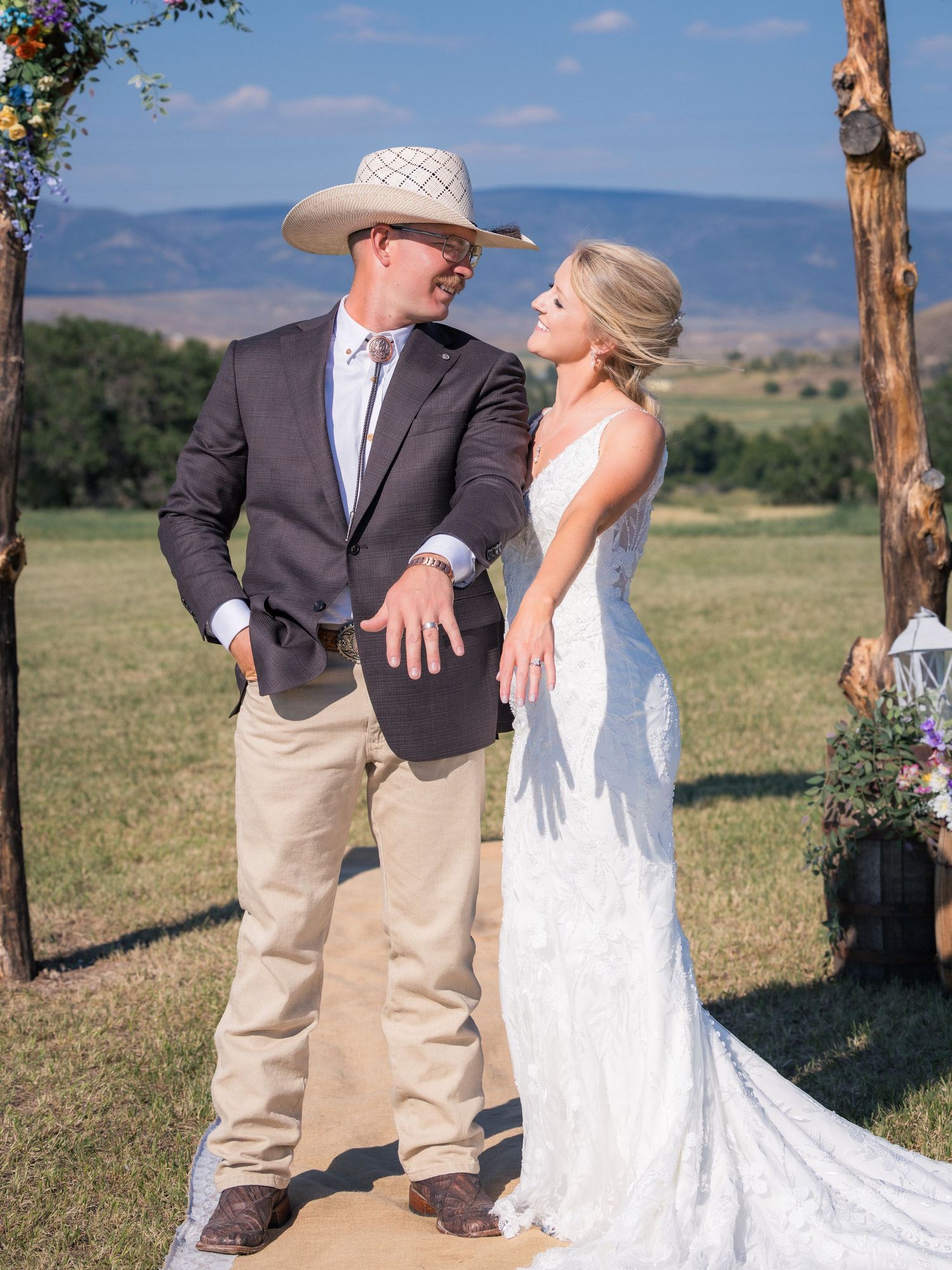 Bride in lace gown and groom in cowboy hat share a romantic moment at an outdoor rustic ranch wedding ceremony.