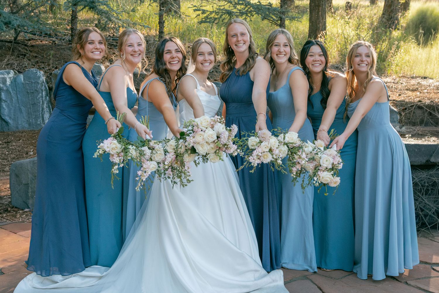 Bride in white gown poses with seven bridesmaids in blue dresses holding floral bouquets outdoors.