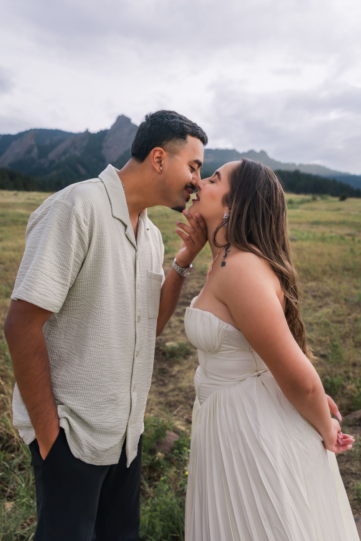 Couple sharing a romantic kiss in a mountain meadow, man in white shirt and woman in white strapless dress.