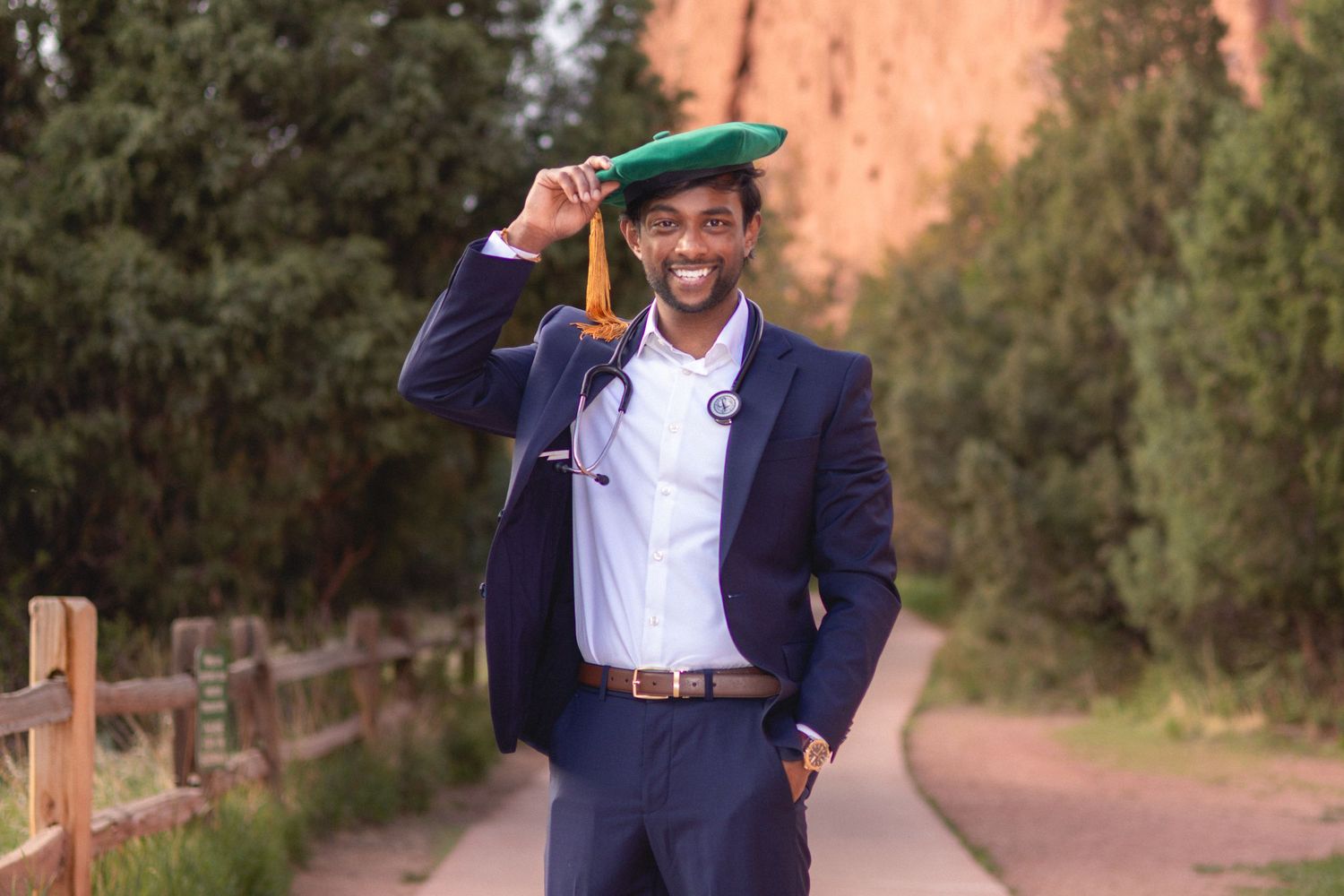 Smiling medical graduate in navy suit holds green cap with stethoscope on scenic outdoor path.