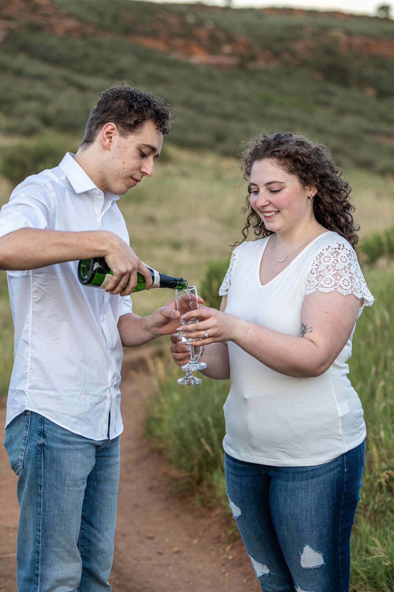 A young couple celebrates outdoors, man pouring champagne into glasses held by smiling woman in white top.